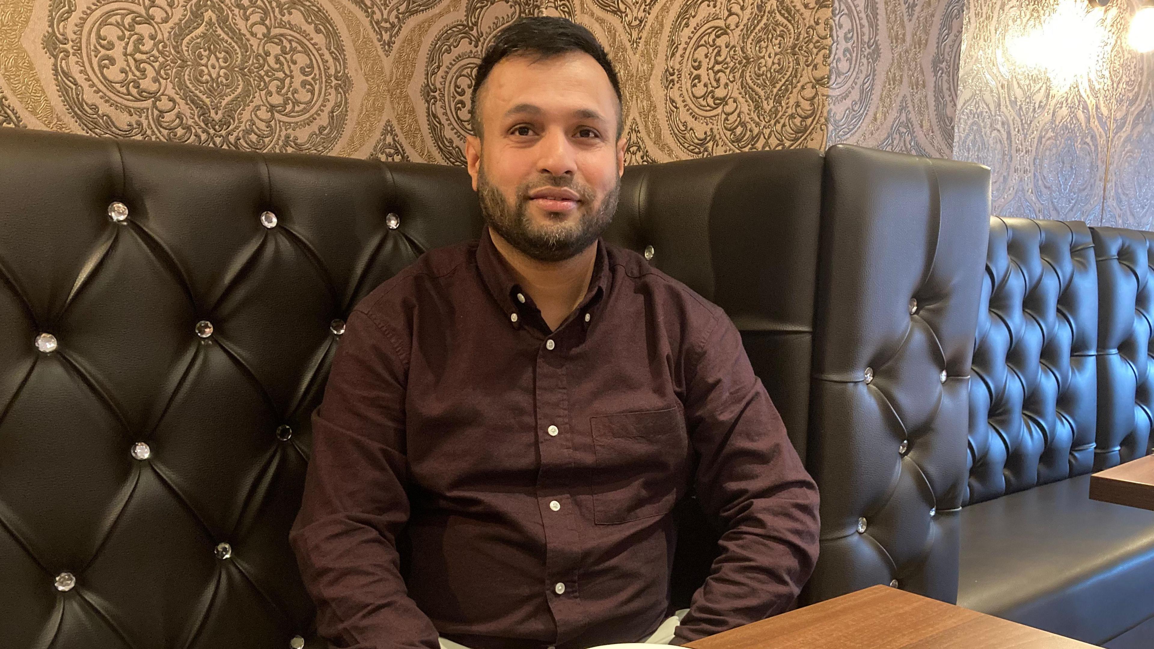 A man wearing a claret coloured buttoned shirt, sat down on a table inside a restaurant - behind him are leather back rests.