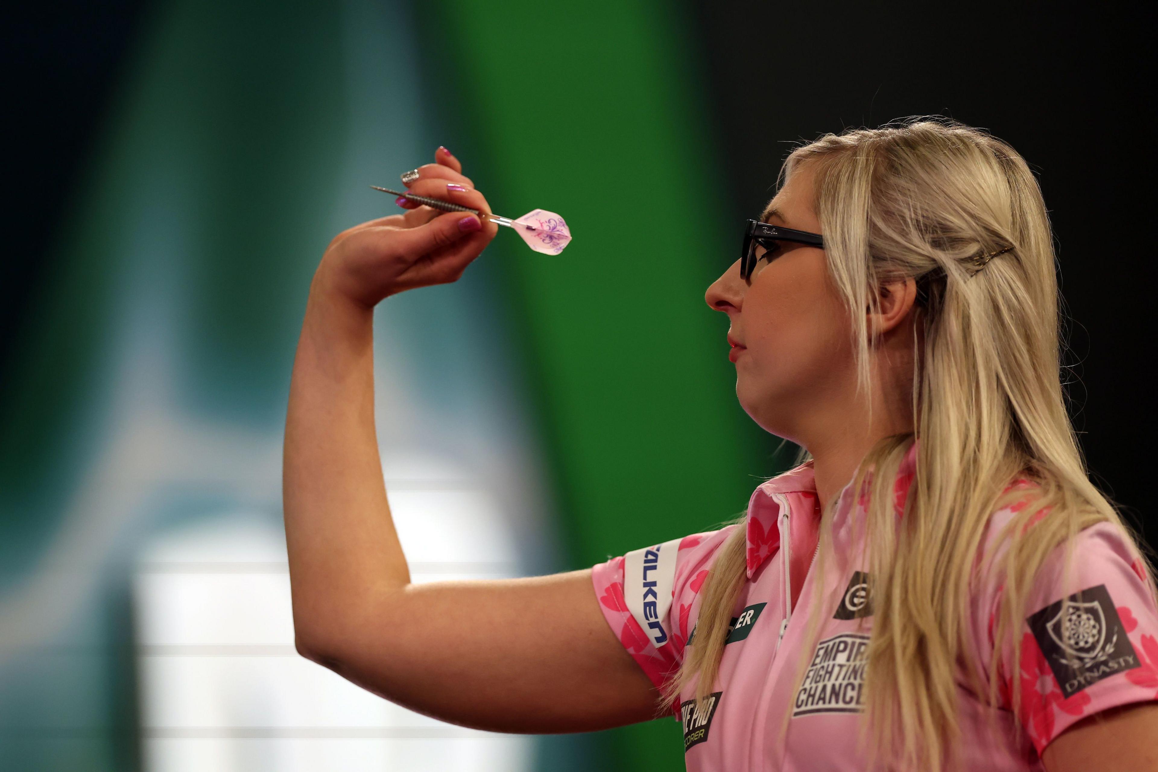 Fallon Sharrock, a blonde woman in glasses and a pink top, throws a dart