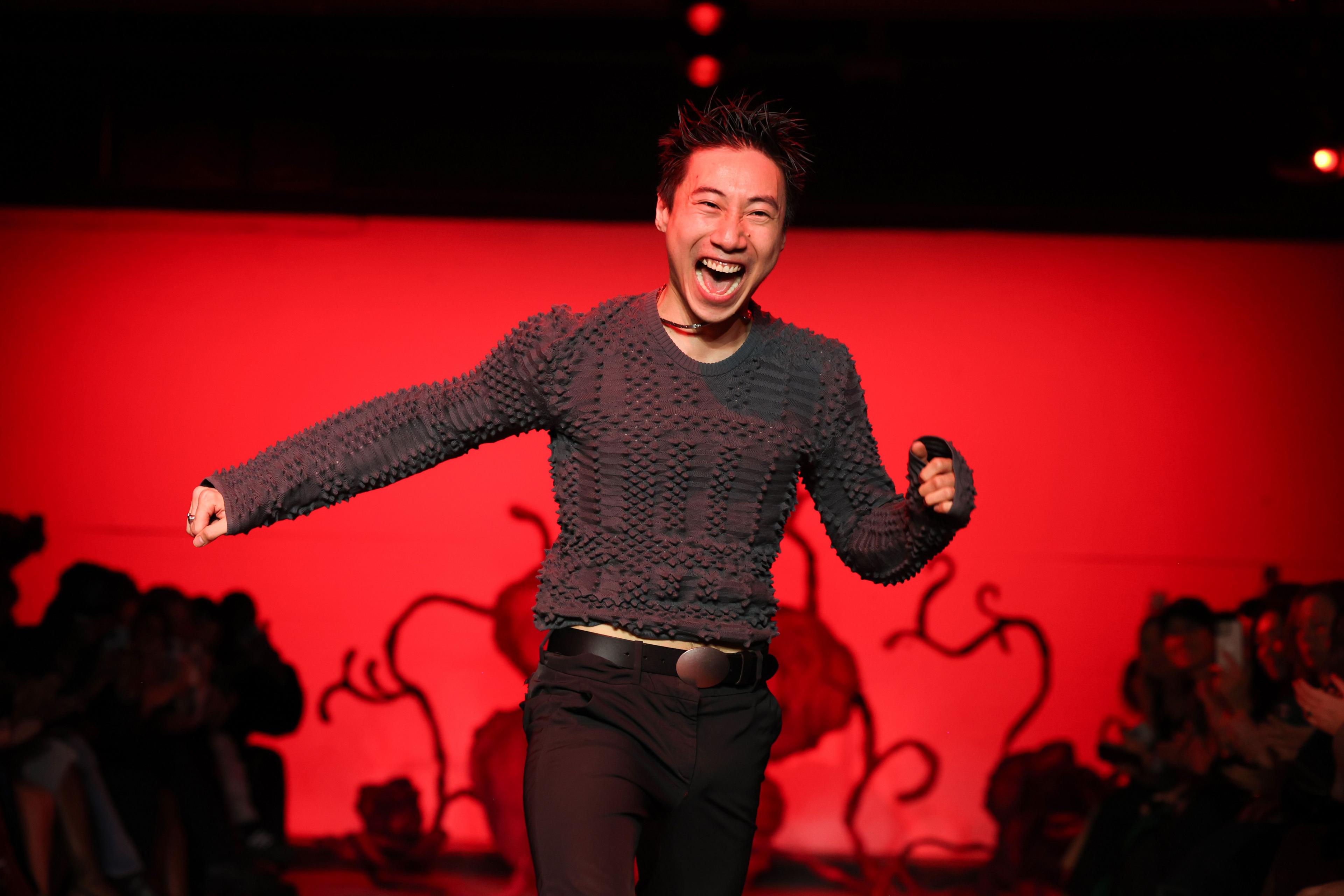 Fashion designer Chet Lo bounding down a catwalk. The background is illuminated red with black squiggles. He wears black trousers and a grey jumper and he is smiling with an open mouth.