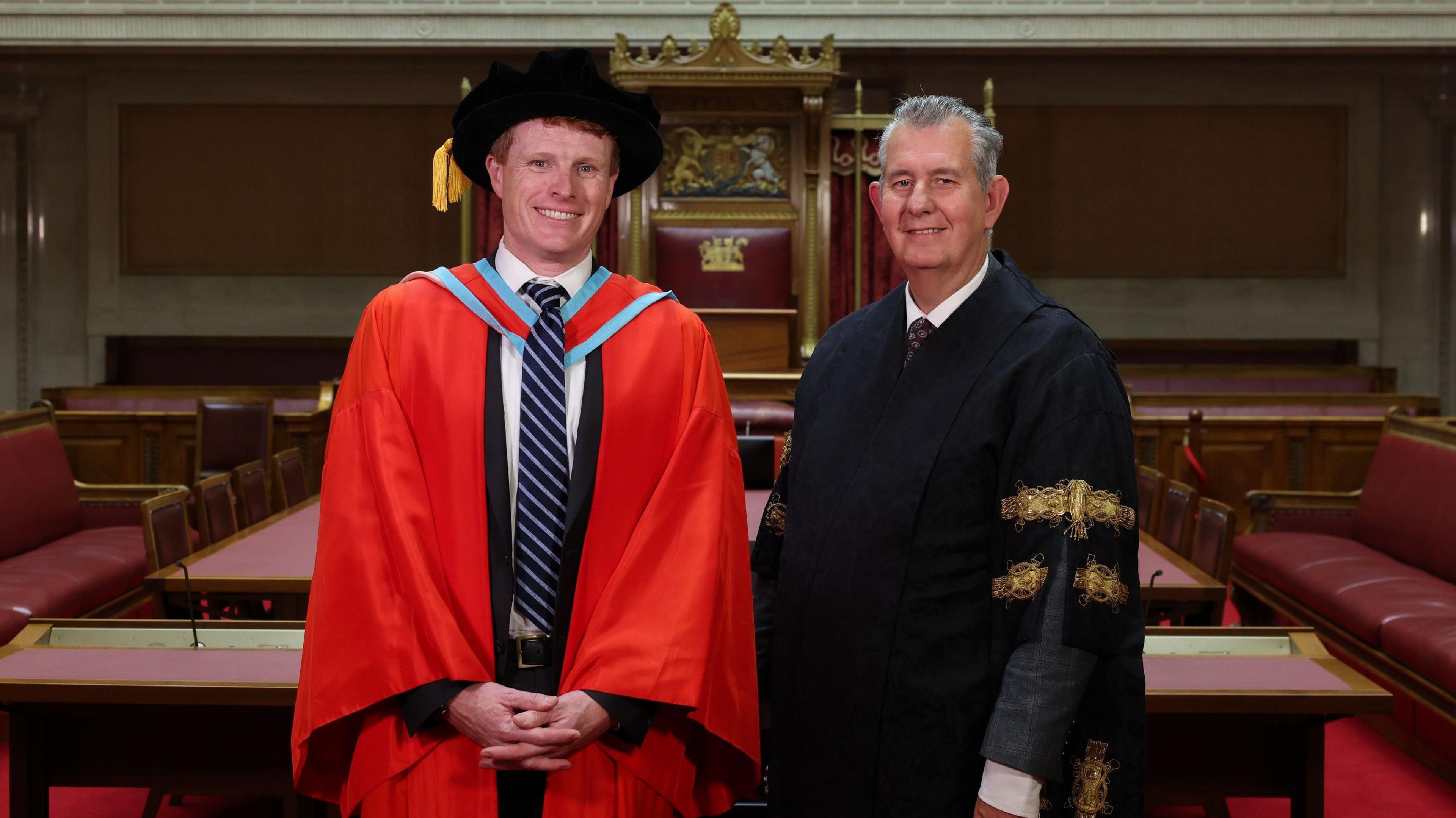 Joe Kennedy standing with has hands clasped in front of him wear red academic robes and a black cap next to the speaker of the Northern Ireland Assembly Edwin Poots who is in a black robe with gold trimming. Behind him the red leather benches of the senate chamber at Stormont. 