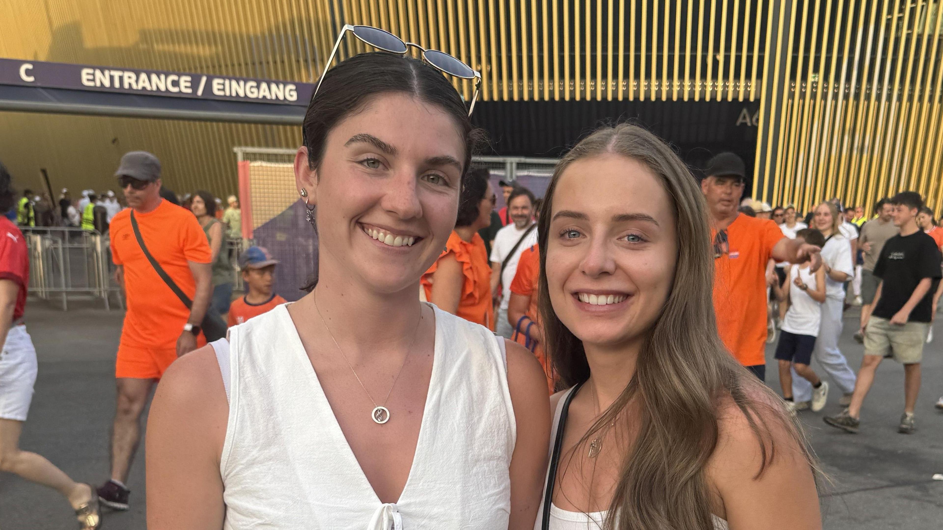 Libby Stott and Nicole Power smiling at the camera in front of the stadium as the fans walk behind them.