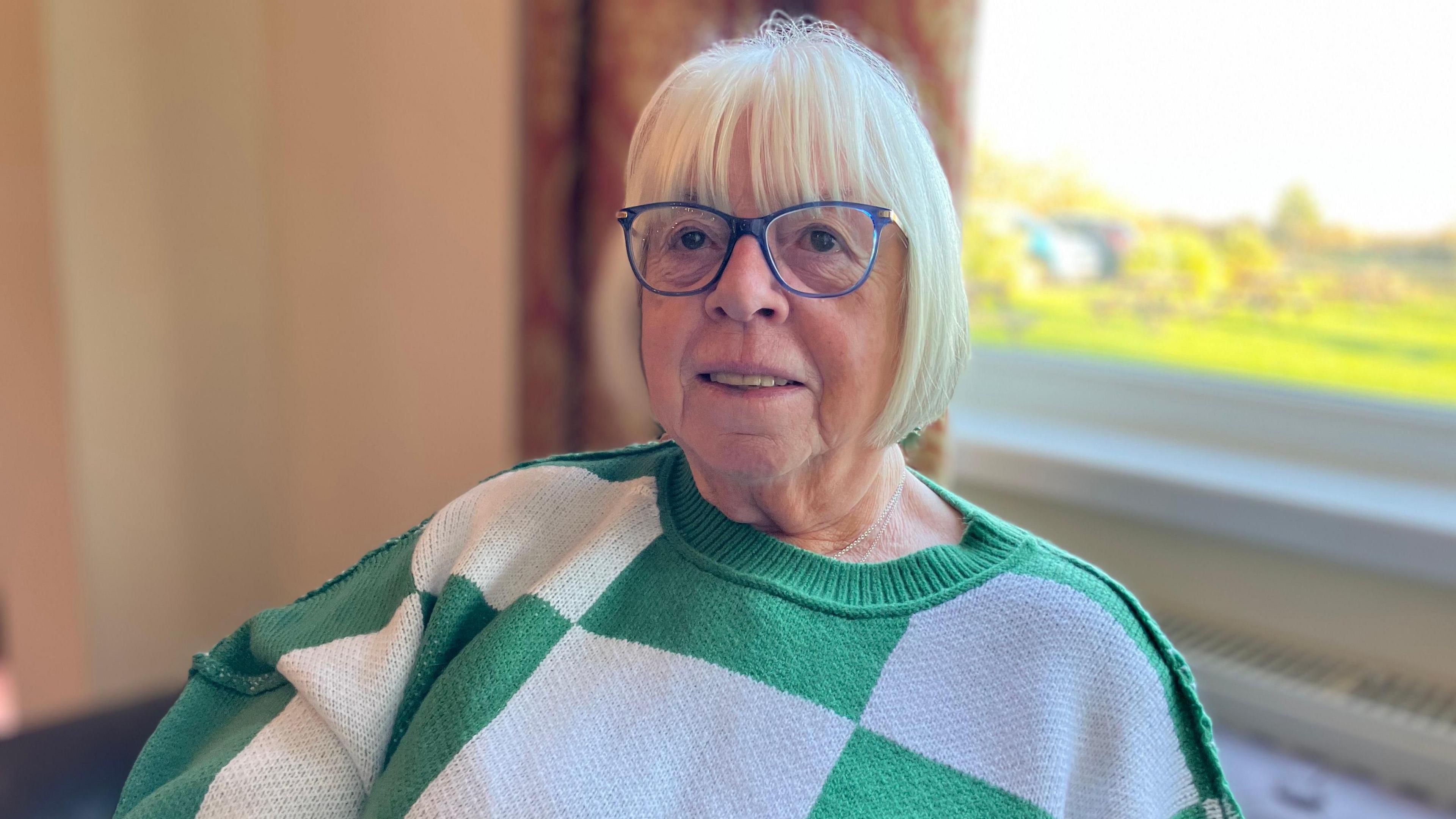 A lady sitting in a chair, She is wearing a green and white jumper and has blue spectacles and fair hair.