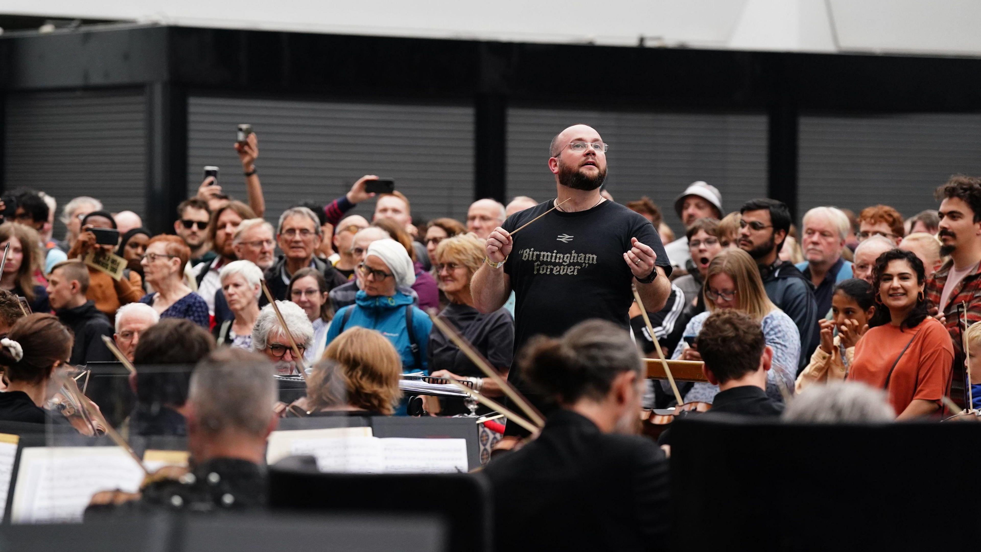 A conductor wearing a black T-shirt that reads "Birmingham forever" leads a large orchestra playing a concert in a station, while a crowd watches behind him.