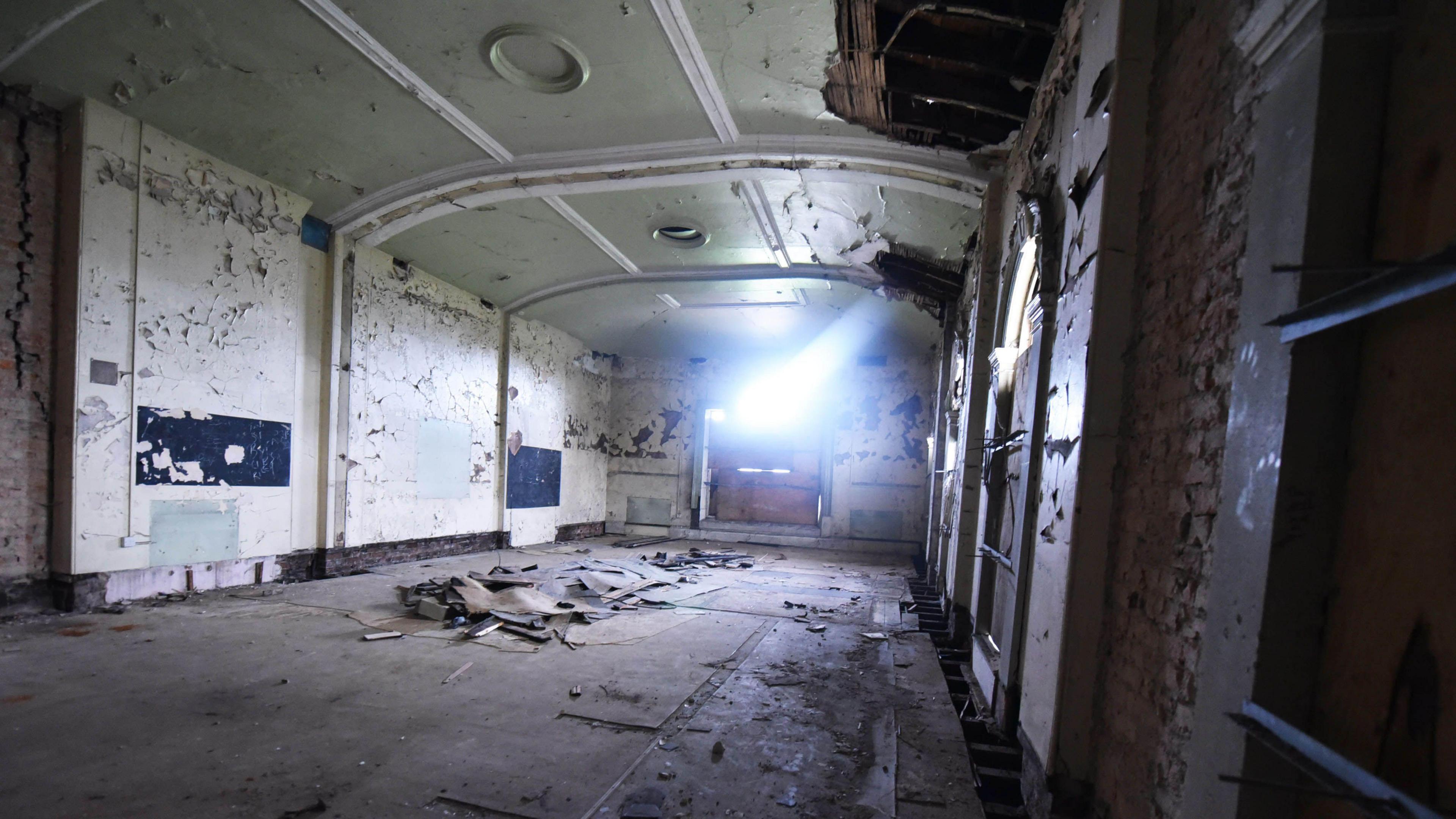 The dilapidated interior of Middlesbrough's Old Town Hall. The room is rectangular-shaped. There are large holes in the ornate ceiling with paint peeling away across each of the walls. The concrete floor is covered with debris. Sunlight is peering through a boarded-up window at the far end of the space.