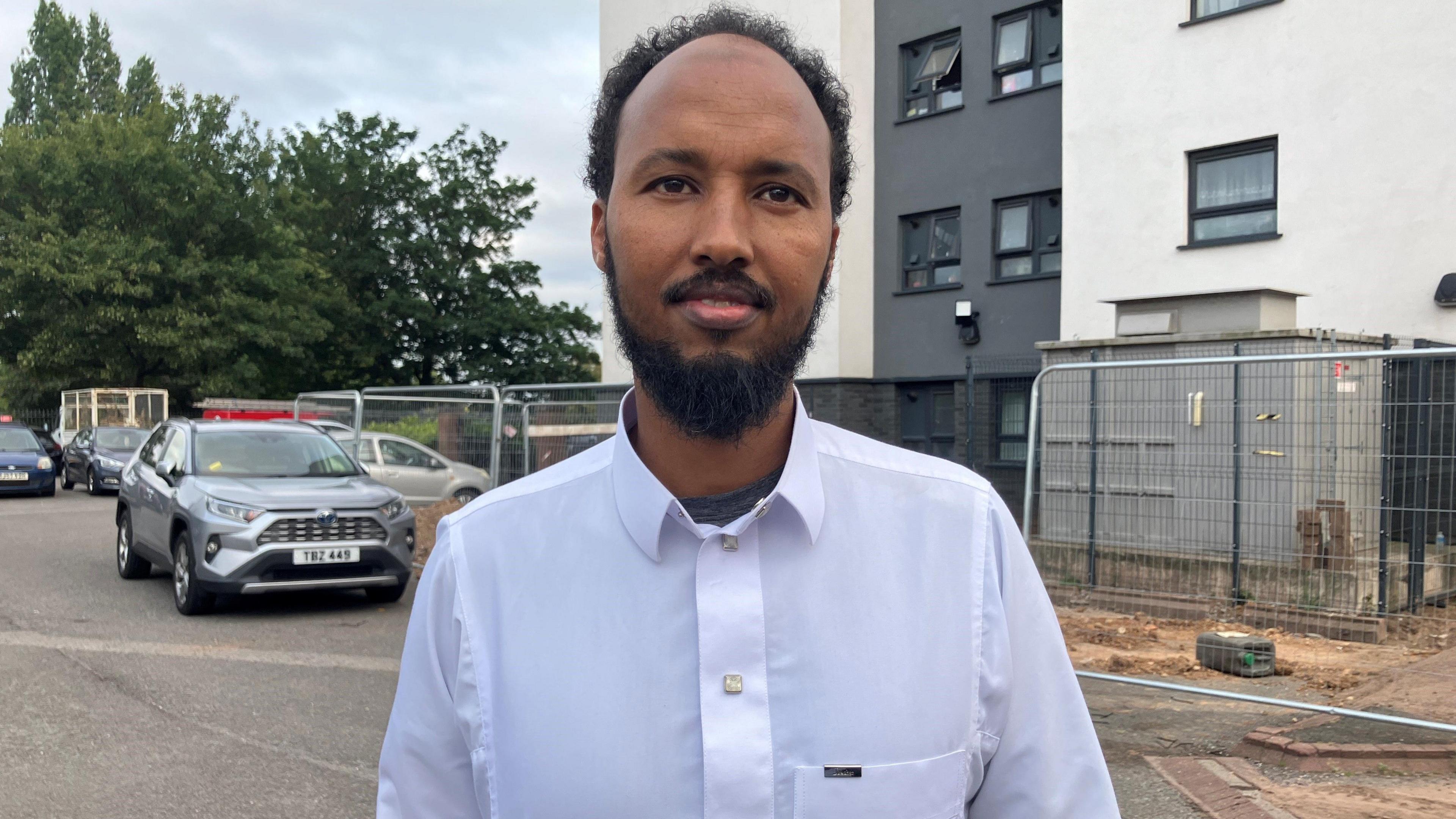 Man looking at camera near to tower block and parked car. He has dark beard and hair, and is wearing a white collared shirt.