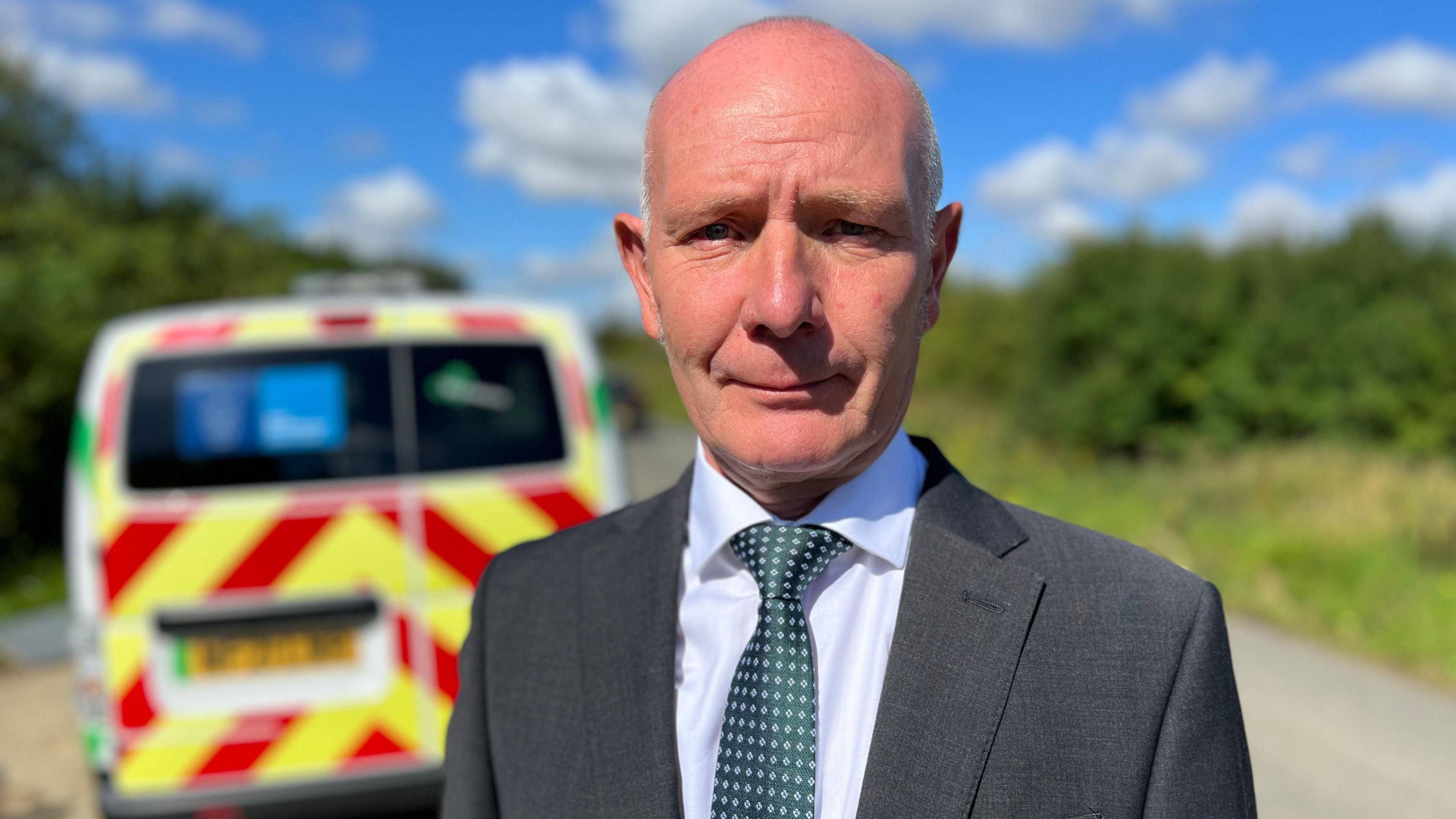Darryl Preston is looking at the camera, standing outside on a sunny day. He is balding and is wearing a grey jacket, white shirt and a green patterned tie. In the background is a blurred image of a police van