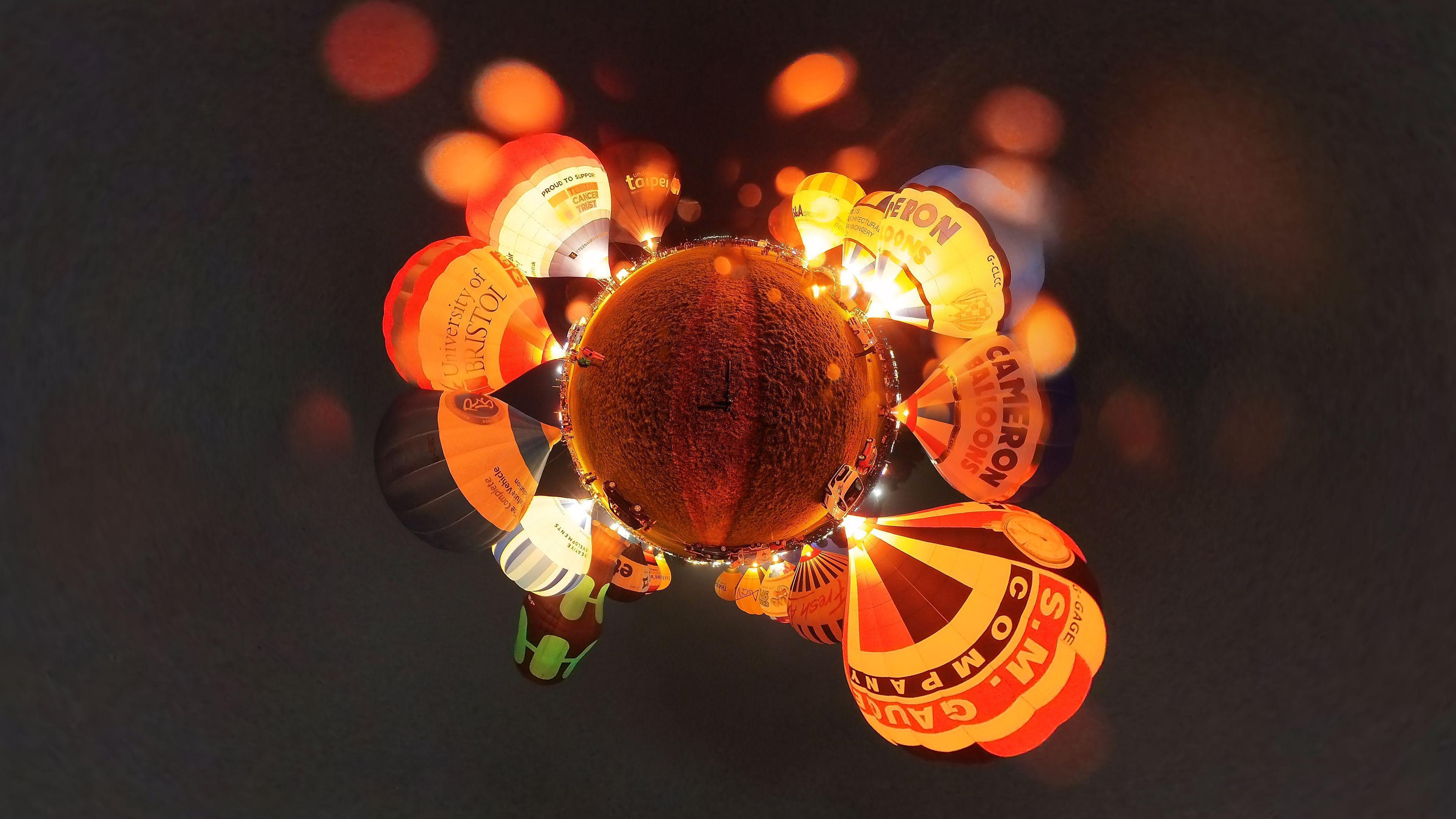 A 360 camera photograph capturing glowing balloons at night, organised in a circle around the ground, which looks like an orb due to the lens