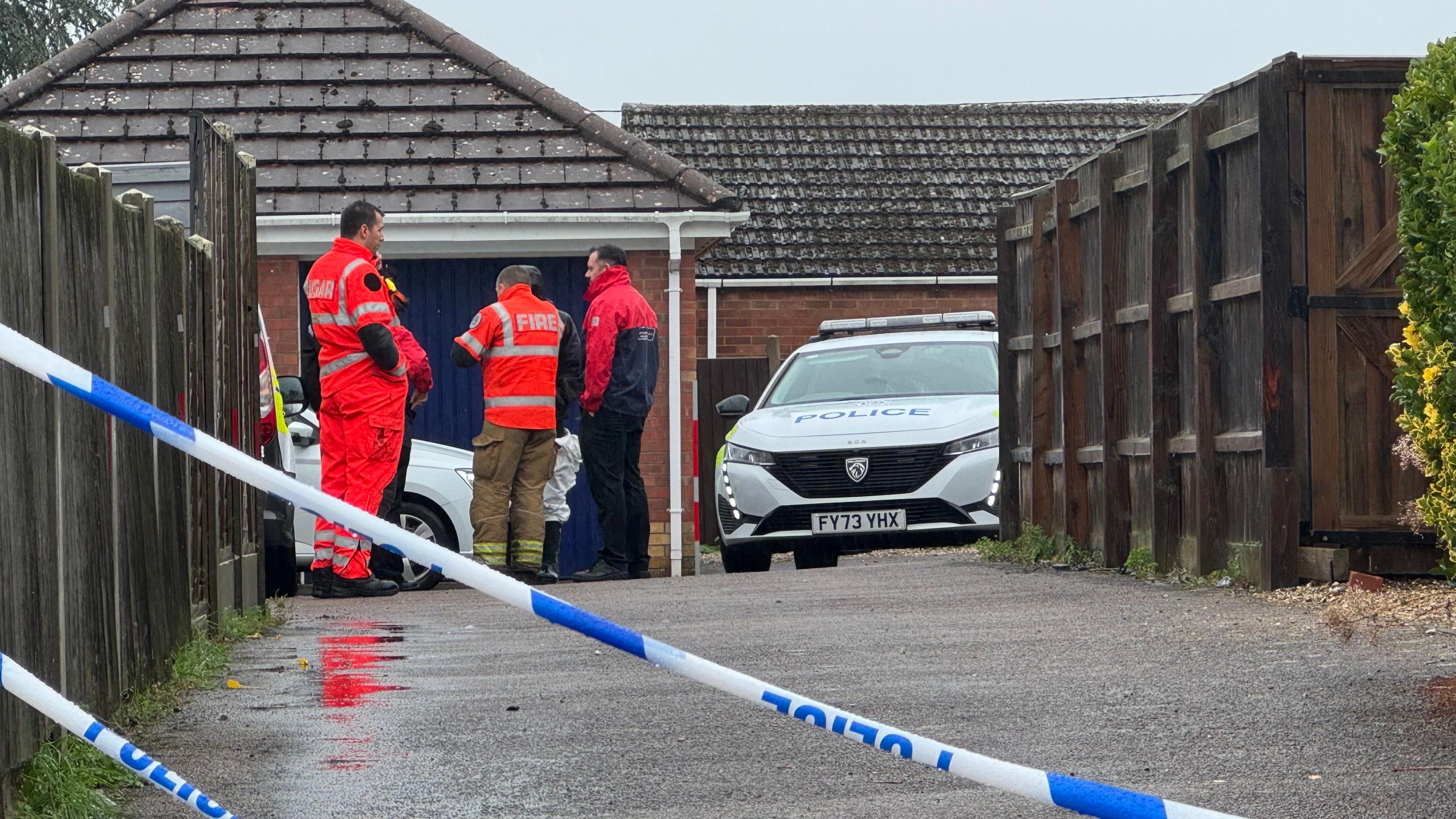 A view down a Tarmac driveway, with wooden fencing either side. At the end of the driveway five figures stand close together. One man is wearing a hi-vis orange jacket with FIRE printed across the back and khaki trousers, another is in hi-vis orange overalls. A police car is parked to the right of the group. Blue and white police tape cuts diagonally across the foreground. The ground is wet from rain.