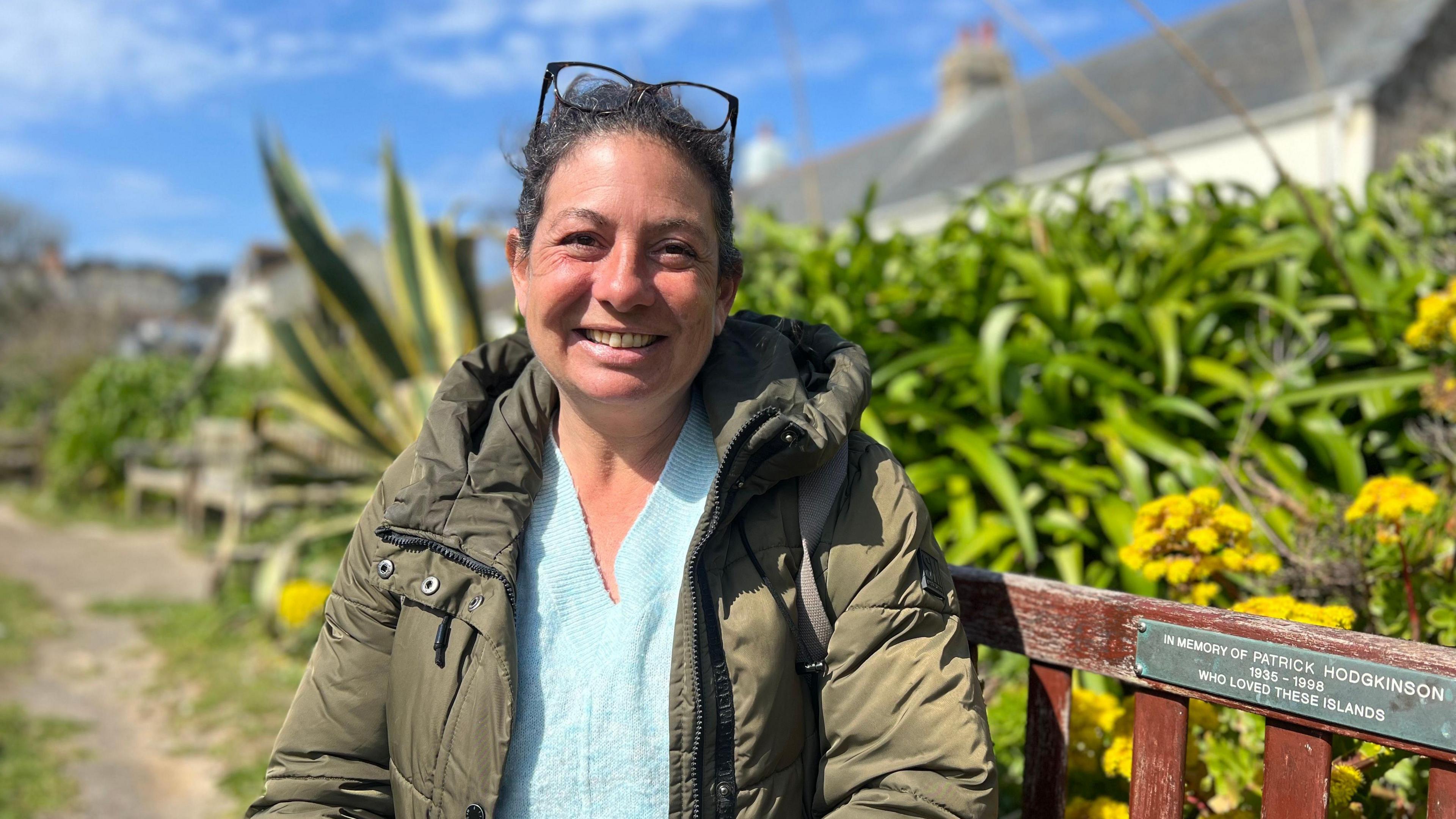Jenny sat on a park bench, she is wearing a light blue jumper and a green coat while smiling at the camera.