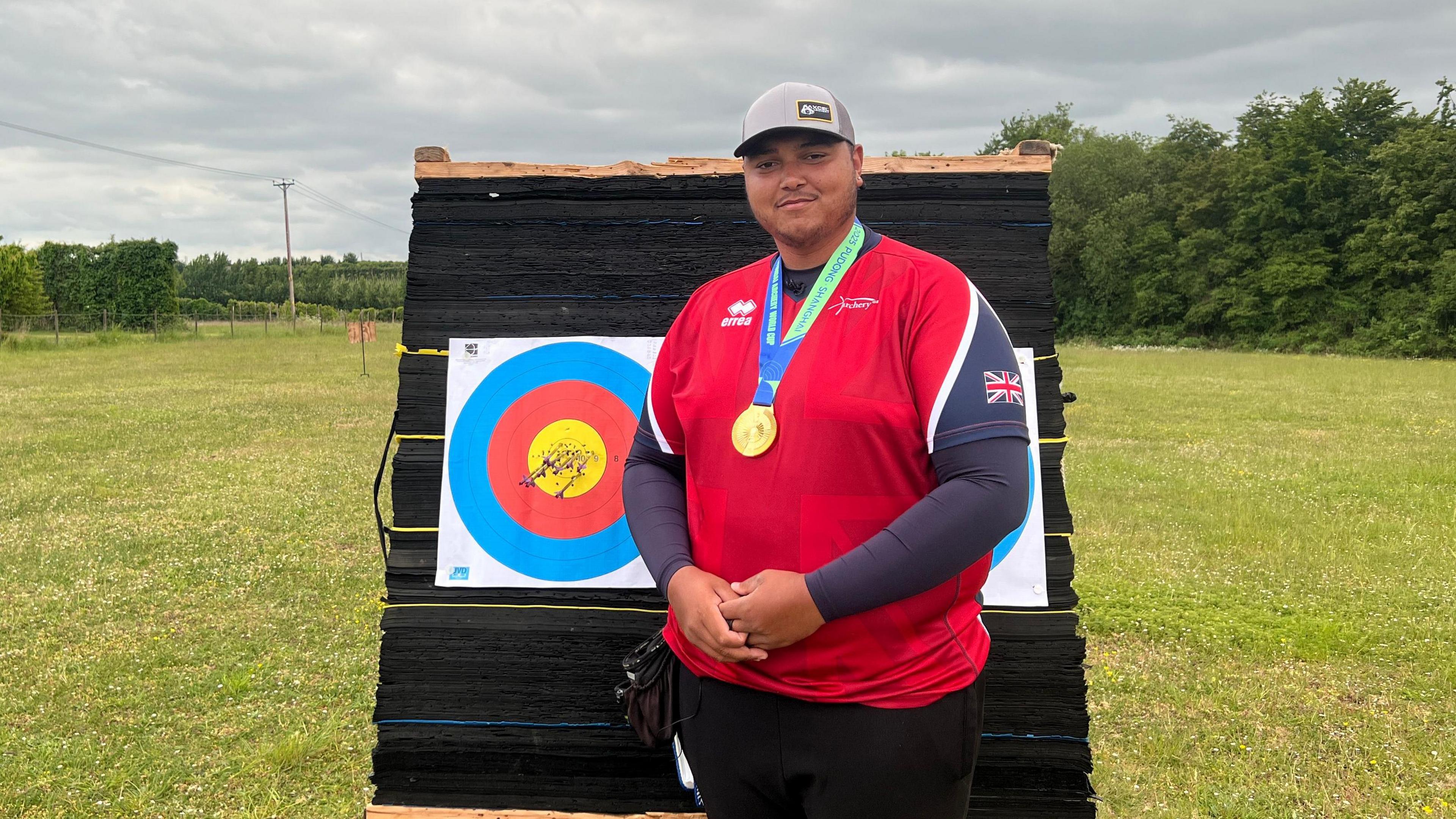 An archer wearing a cap, GB kit and gold medal stands in front of a target looking satisfied. A group of arrows have been shot directly into the centre of the target