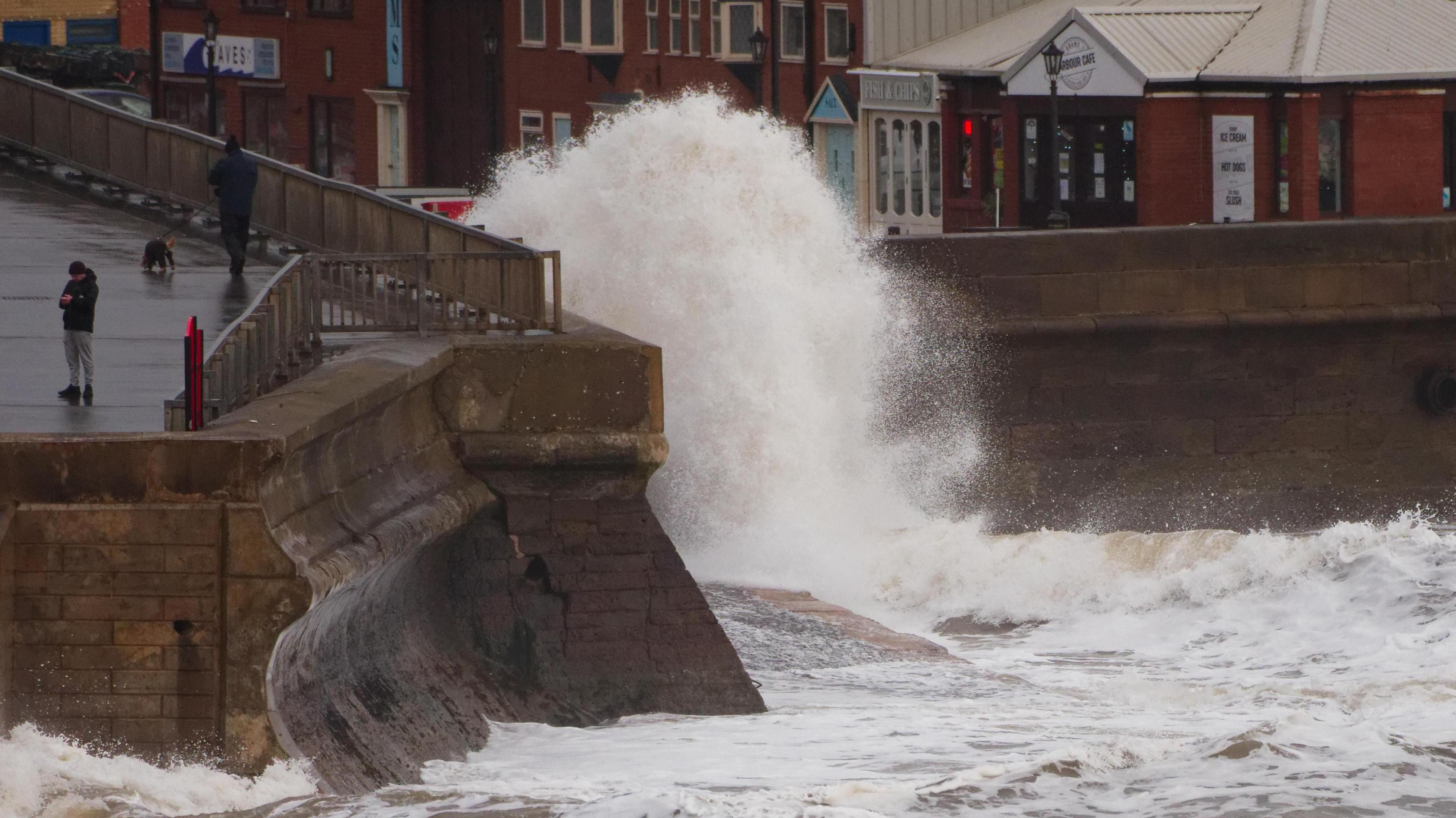 A sea front with tall waves crashing against a stone wall and railings. A figure is walking a dog on the left side of the image. 