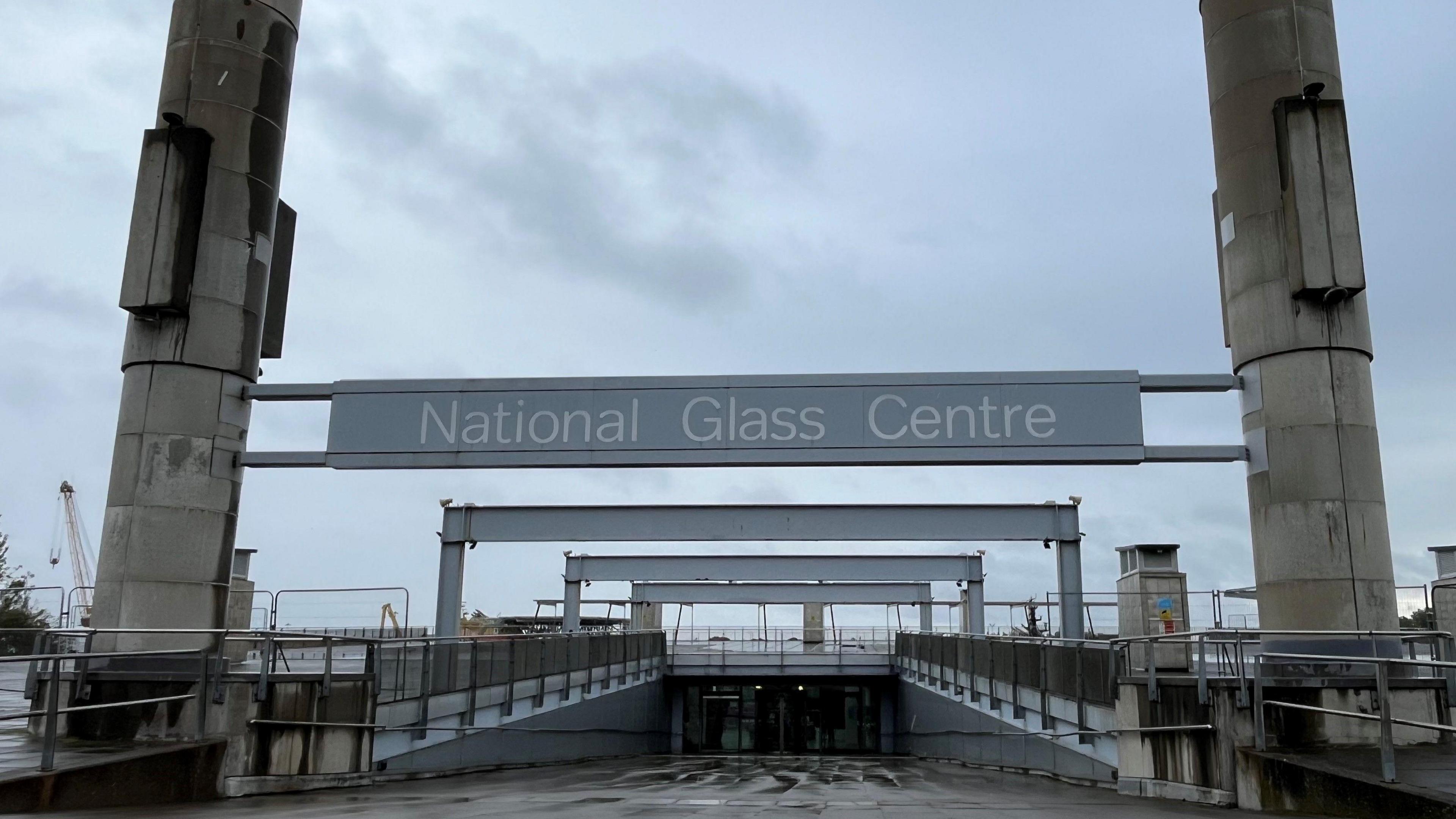 The entrance of the National Glass Centre in Sunderland. A large grey sign is suspended between two high cement pillars. A sloped path leads to the glass doors of the building. The fenced off roof of the building is visible in the distance.