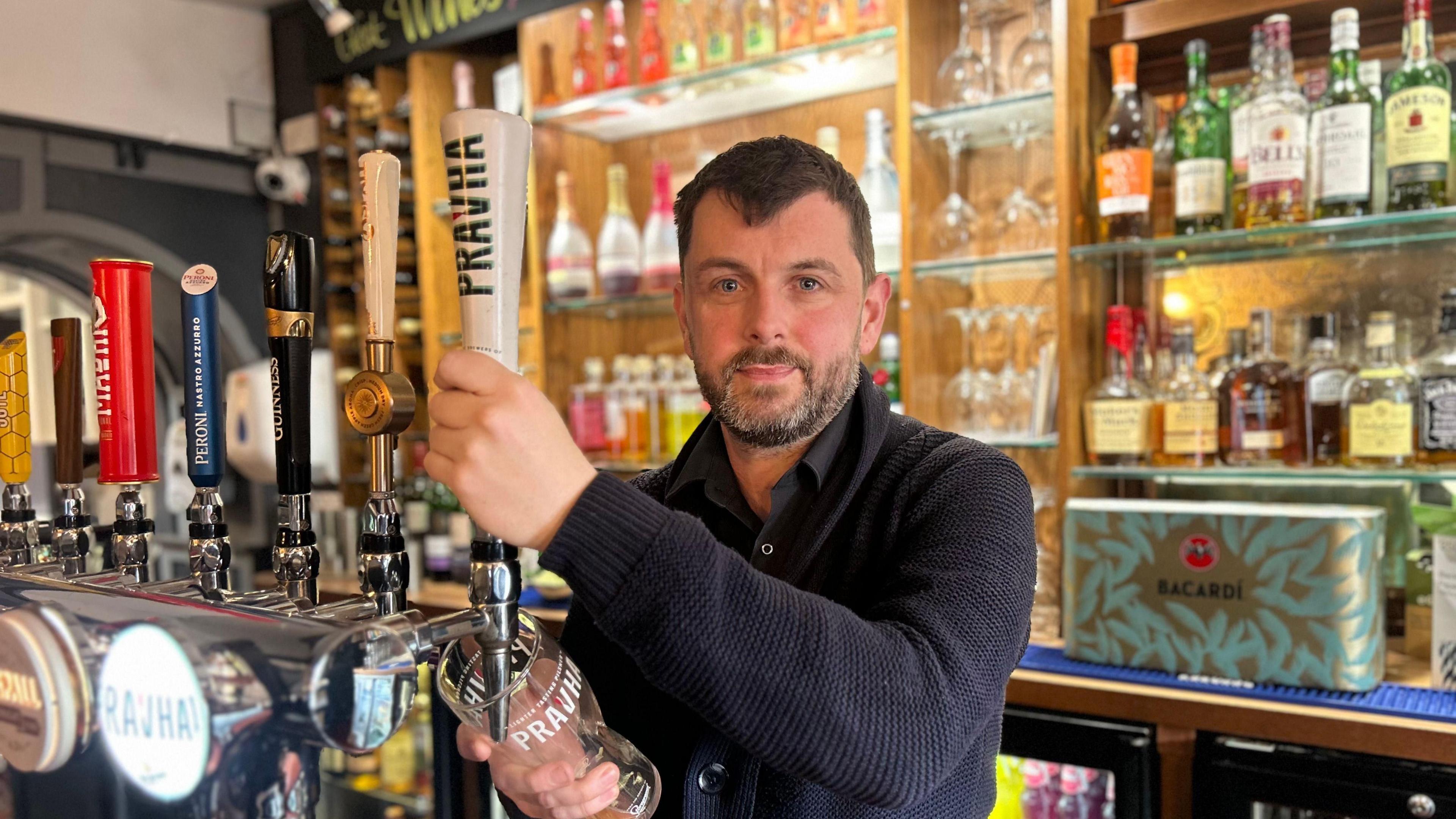 Shot of Allen Slinger pulling a pint behind the brightly-lit bar at the Ferguson Fawsitt Arms. He has dark hair and a beard and is wearing a blue jumper.