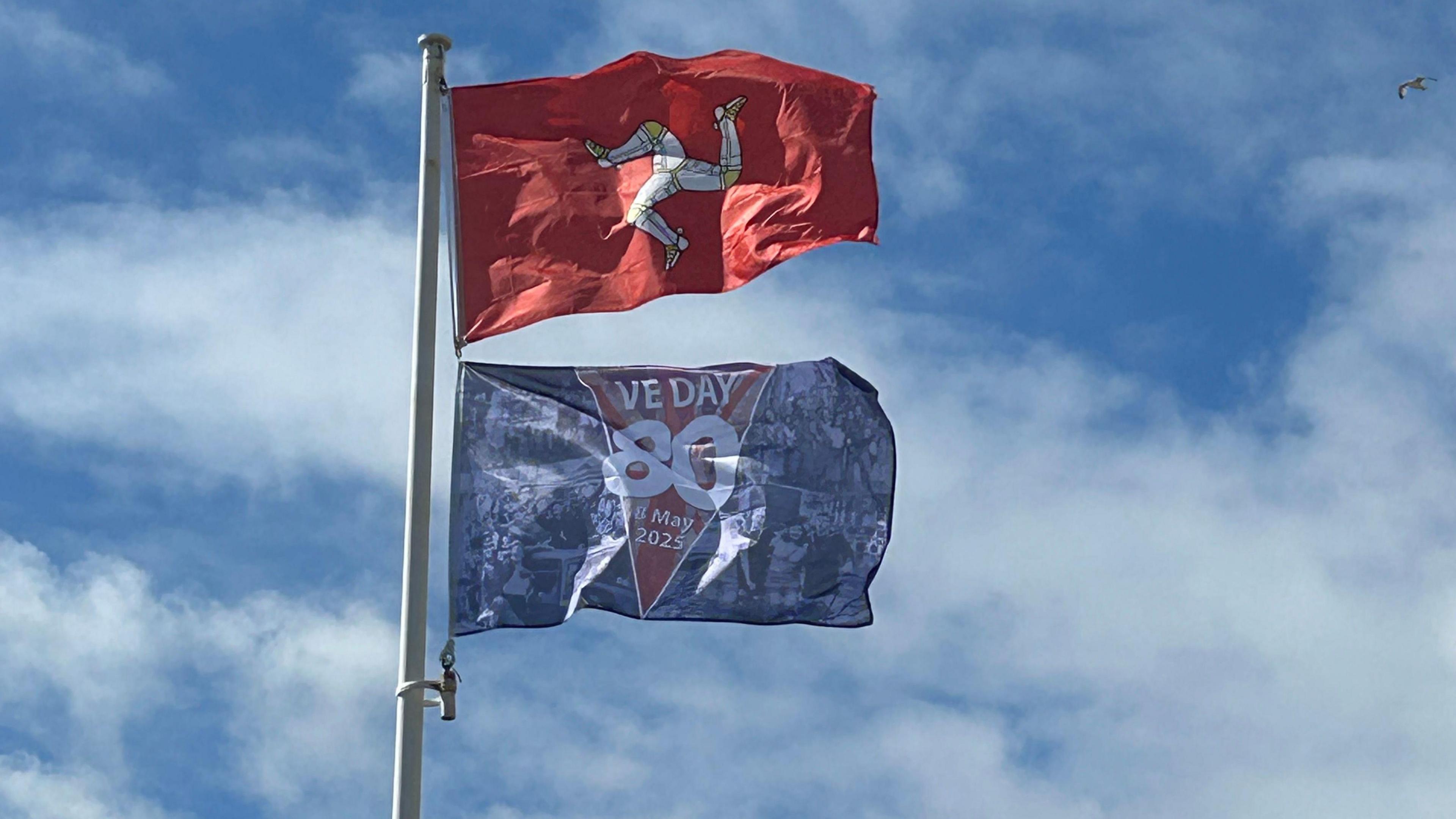 A red Manx flag with a yellow and white triskllion flies on a flag pole. Underneath there is a blue VE Day 80 flag. The sky behind is blue with white clouds.