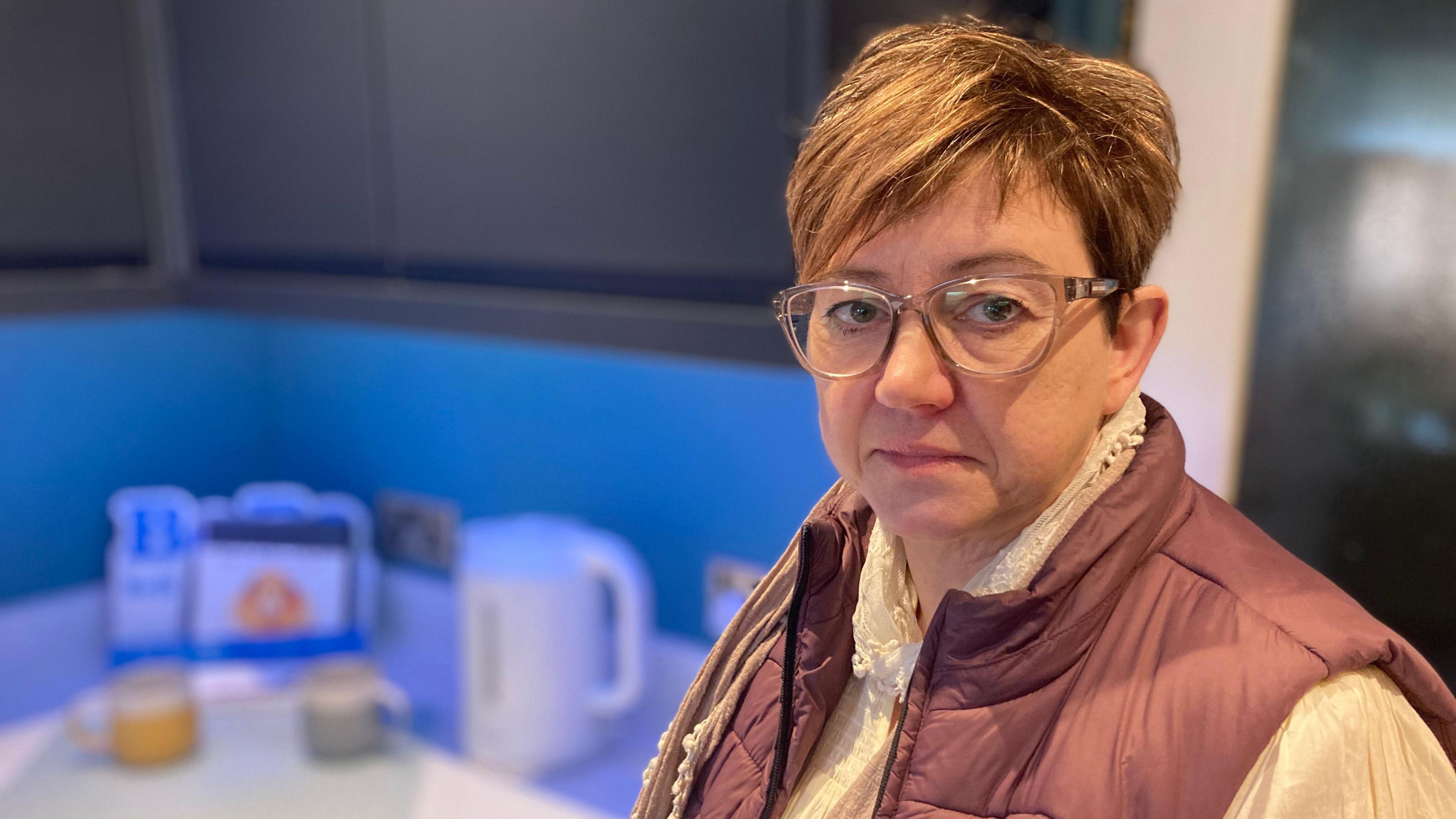 Joanne Davey, a middle aged woman with cropped brown hair and glasses, wearing a brown gilet and cream blouse, looks towards the camera. She is standing in her kitchen with a kettle visible in the background.