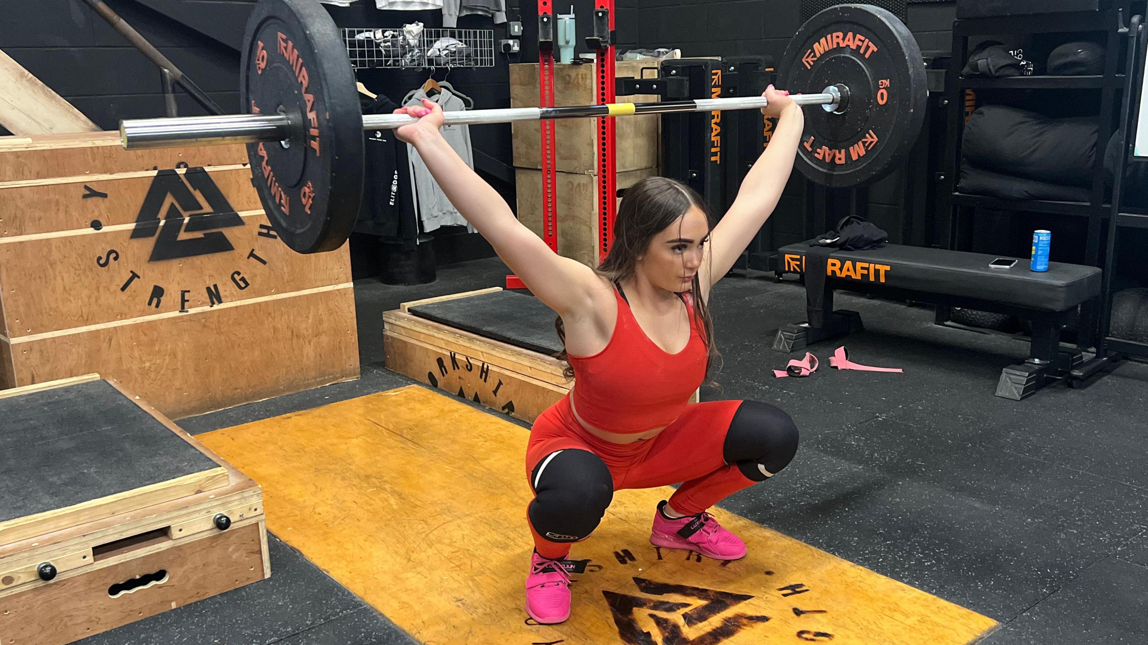 Annie in a squat position, holding a weighted barbell over her head. She is wearing a red gym set and training for the IWF Youth World Championships.