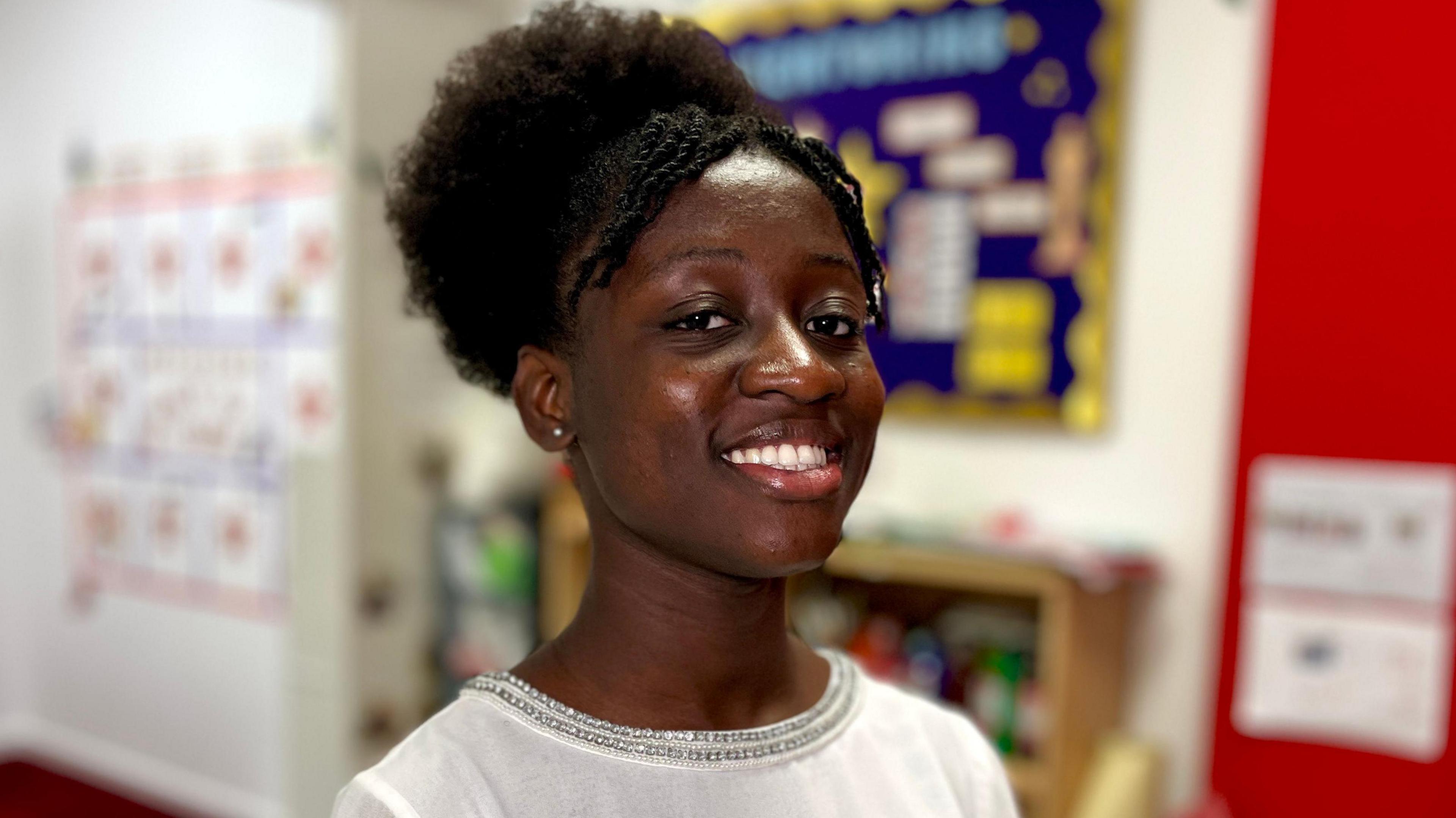 Olivia Ojieman stands in a school setting with a red wall and a bookcase behind her. She has medium-length black hair tied back is smiling at the camera. She is wearing a white top with a silver trim.