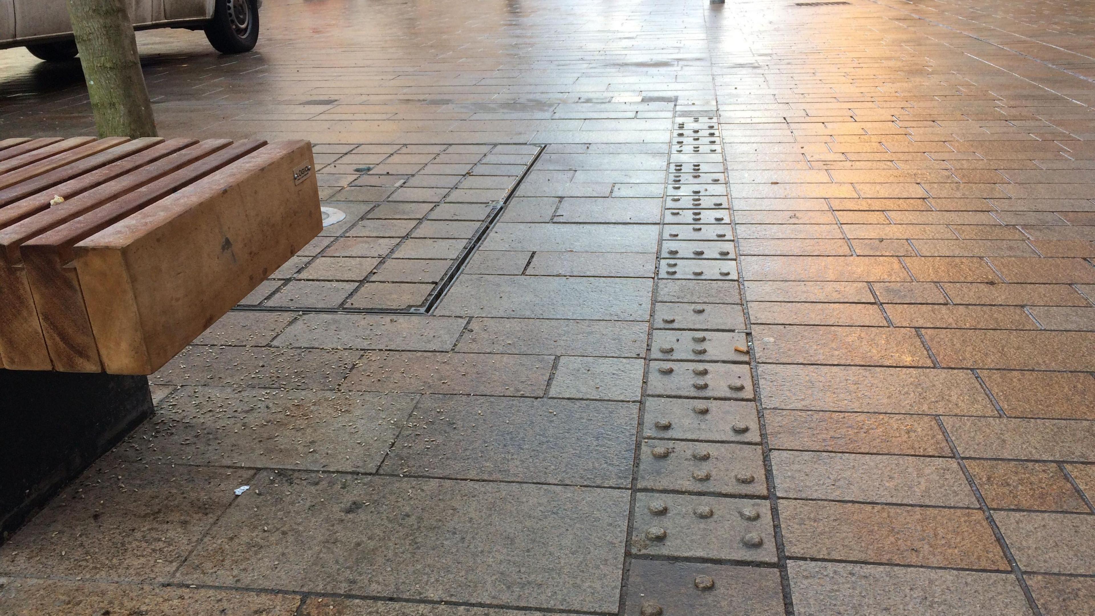 Street paving with a row of slabs with Braille raised markings in a straight line and a wooden bench to the left.