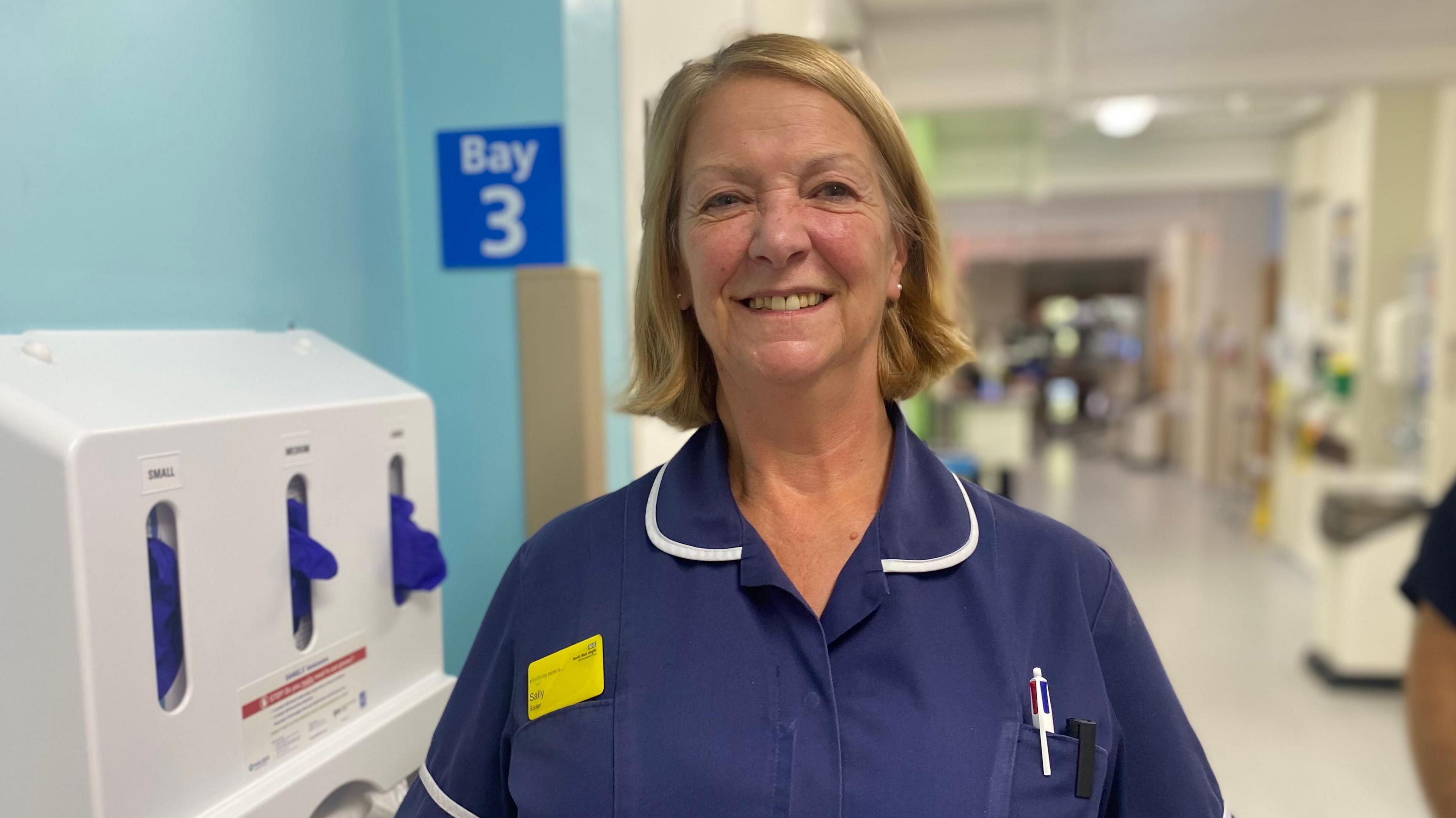 Sally Cotton is smiling at the camera while standing in a hospital ward. She is wearing a navy blue top and has a short, fair bob. She has a yellow name badge on and there are pens in her top pocket