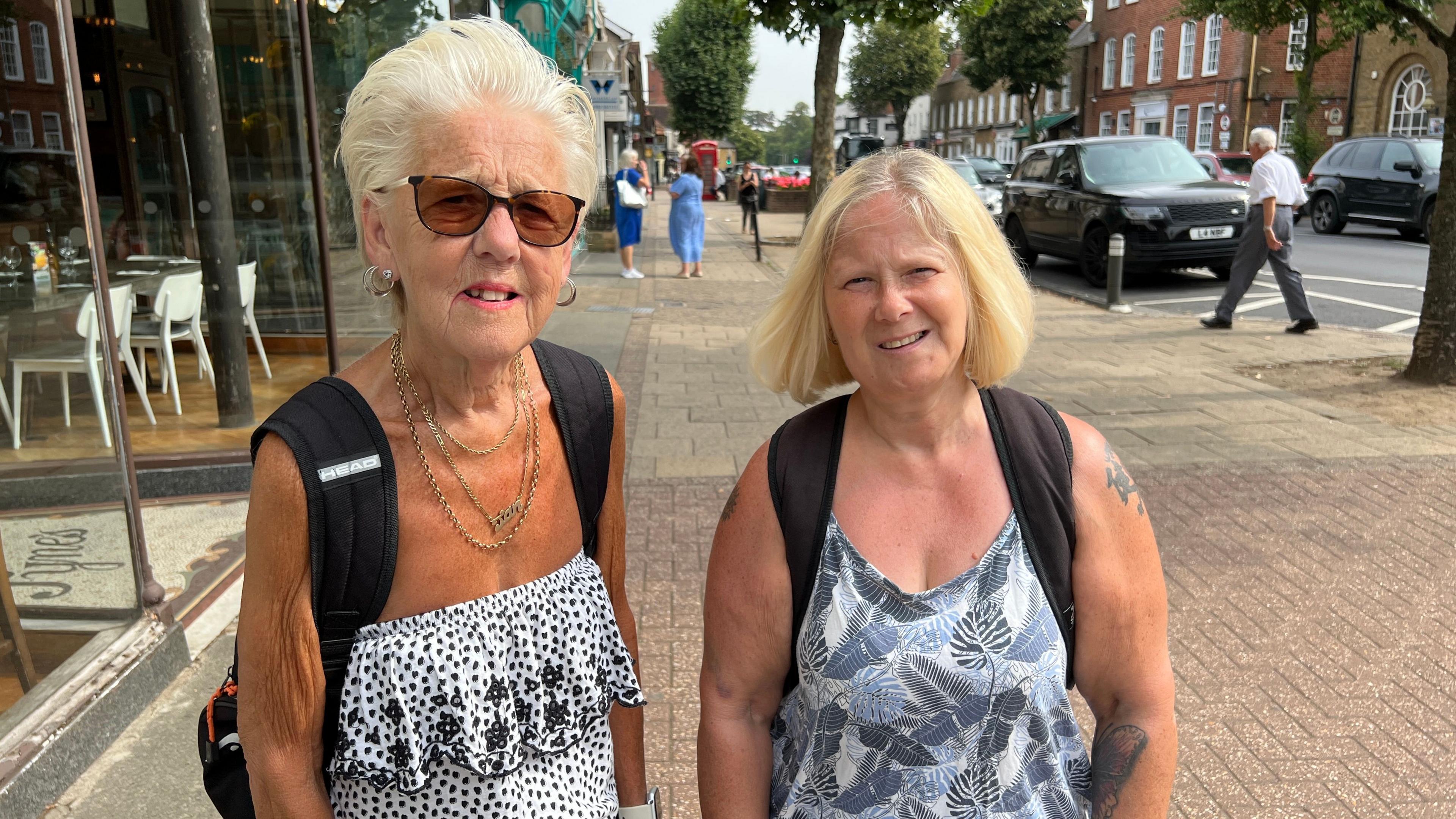 Two women wearing back packs stand in a street looking at the camera.