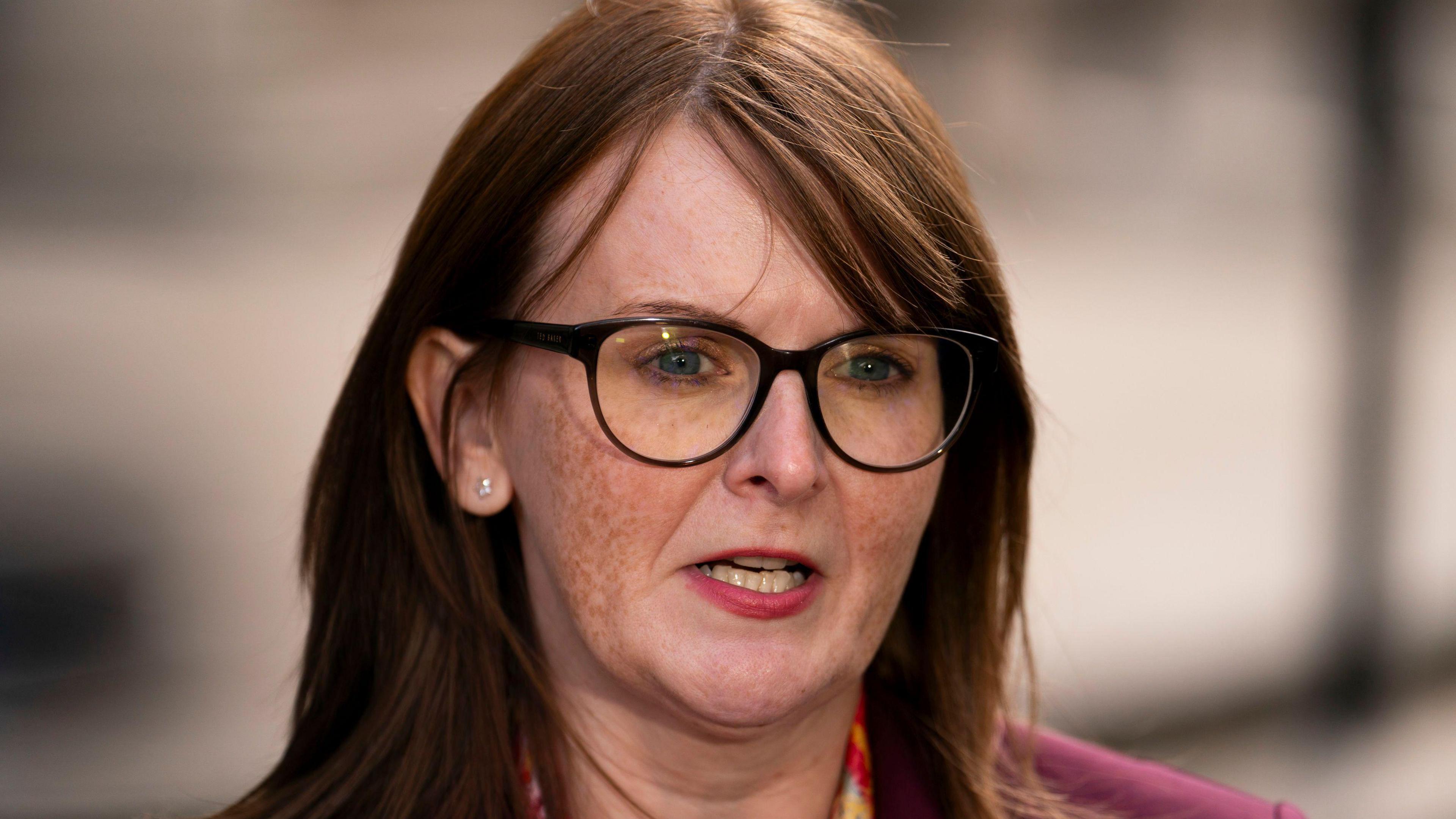 Caoimhe Archibald with brown/copper long hair and a side fringe. She is wearing dark framed glasses, a diamond stud earring in the ear not covered by her hair. She is mid-speech outside the Treasury in London. She is wearing a pink jacket and pink, red, yellow and orange floral scarf. 