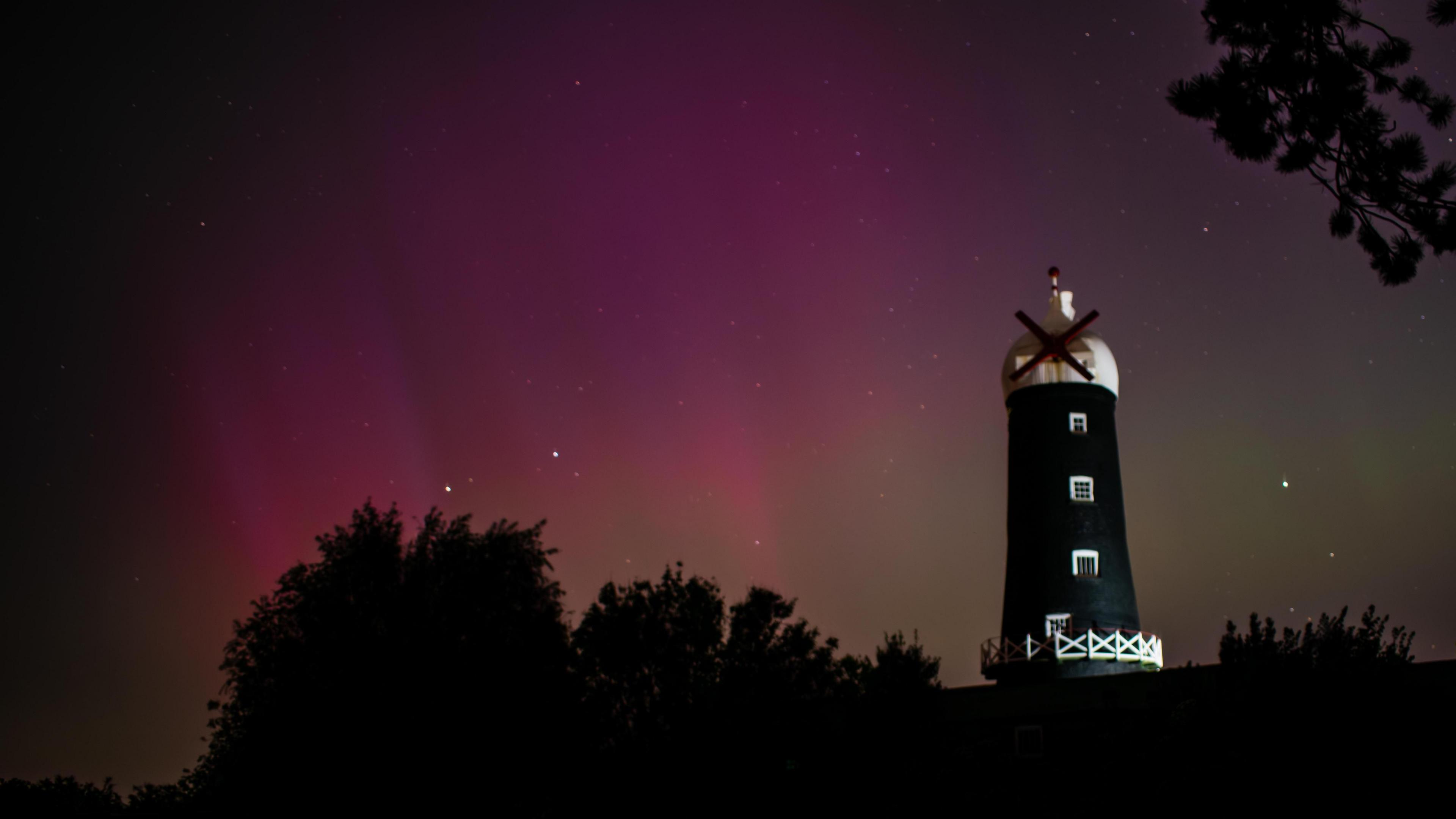 A photo showing the pink hues of the Northern Lights, with a large black and white windmill in the shot.