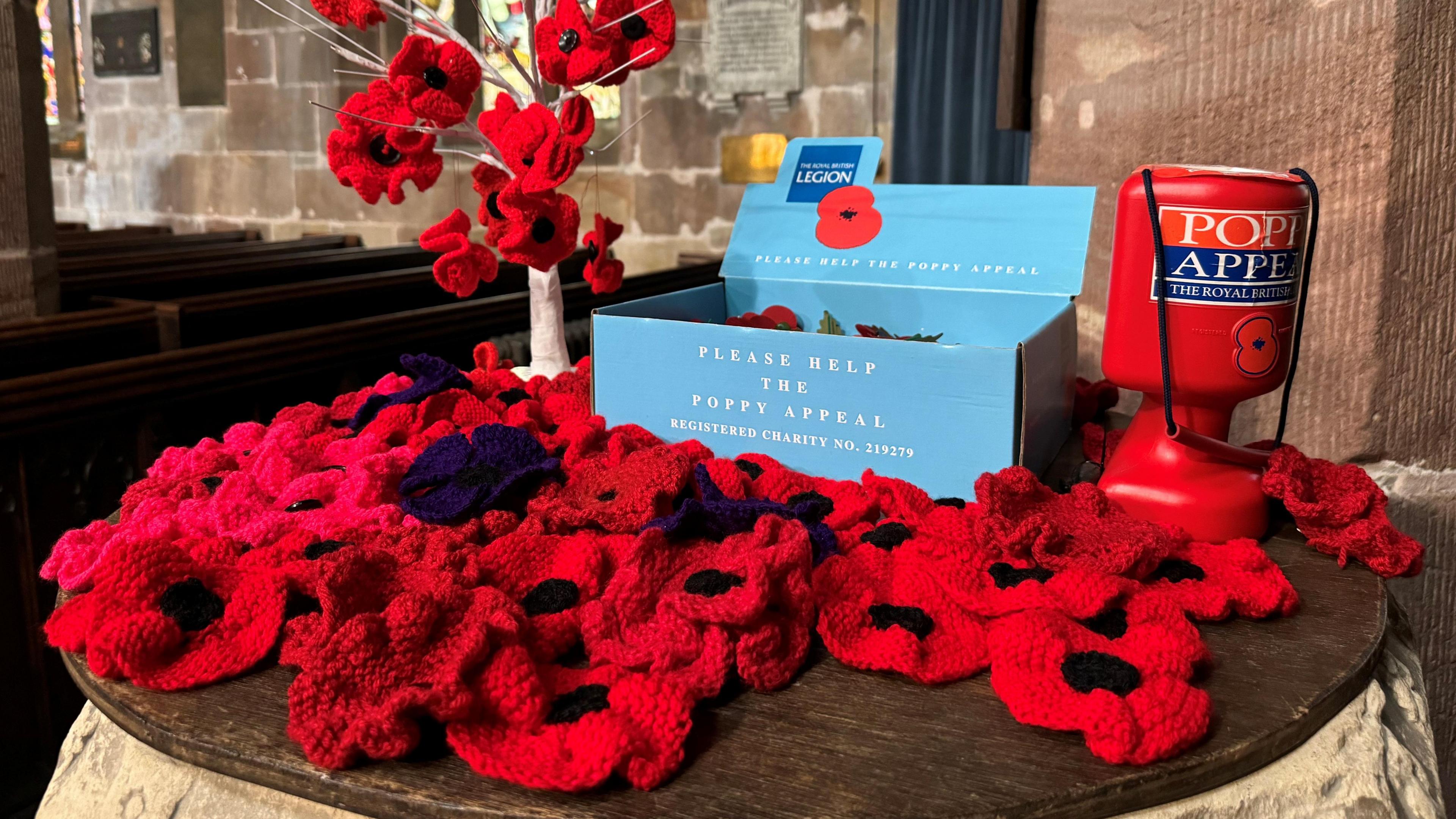 A Royal British Legion Poppy Appeal collection tin sits next to a blue box which contains paper poppies, both are are surrounded by knitted poppies sat on a wooden surface