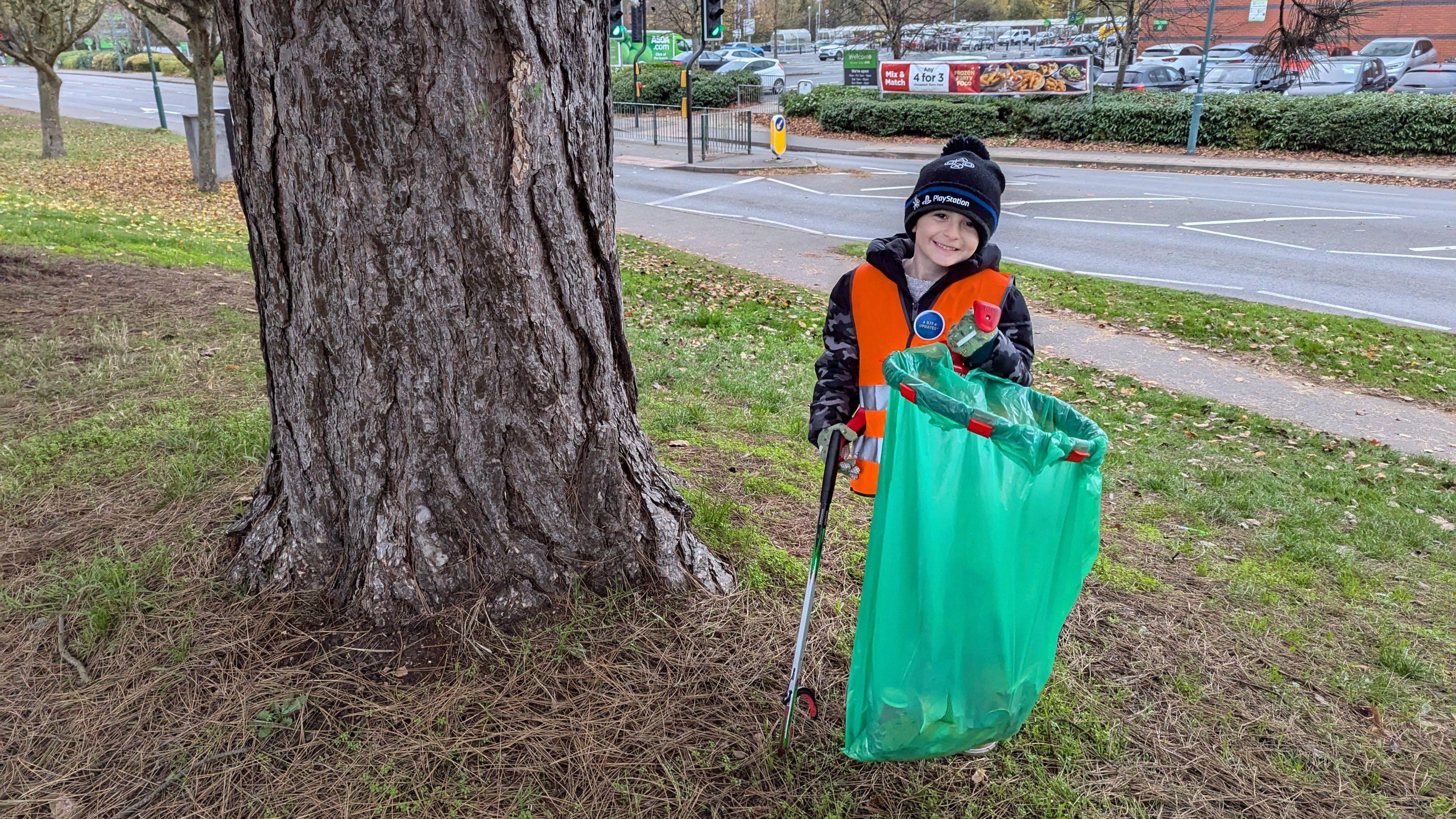 Harlee is standing next to a large tree on some grass. He is wearing a black and dark blue hat, with a grey and black camo coat, and an orange hi-vis jacket. He is holding a green waste bag and a metal litter picking tool