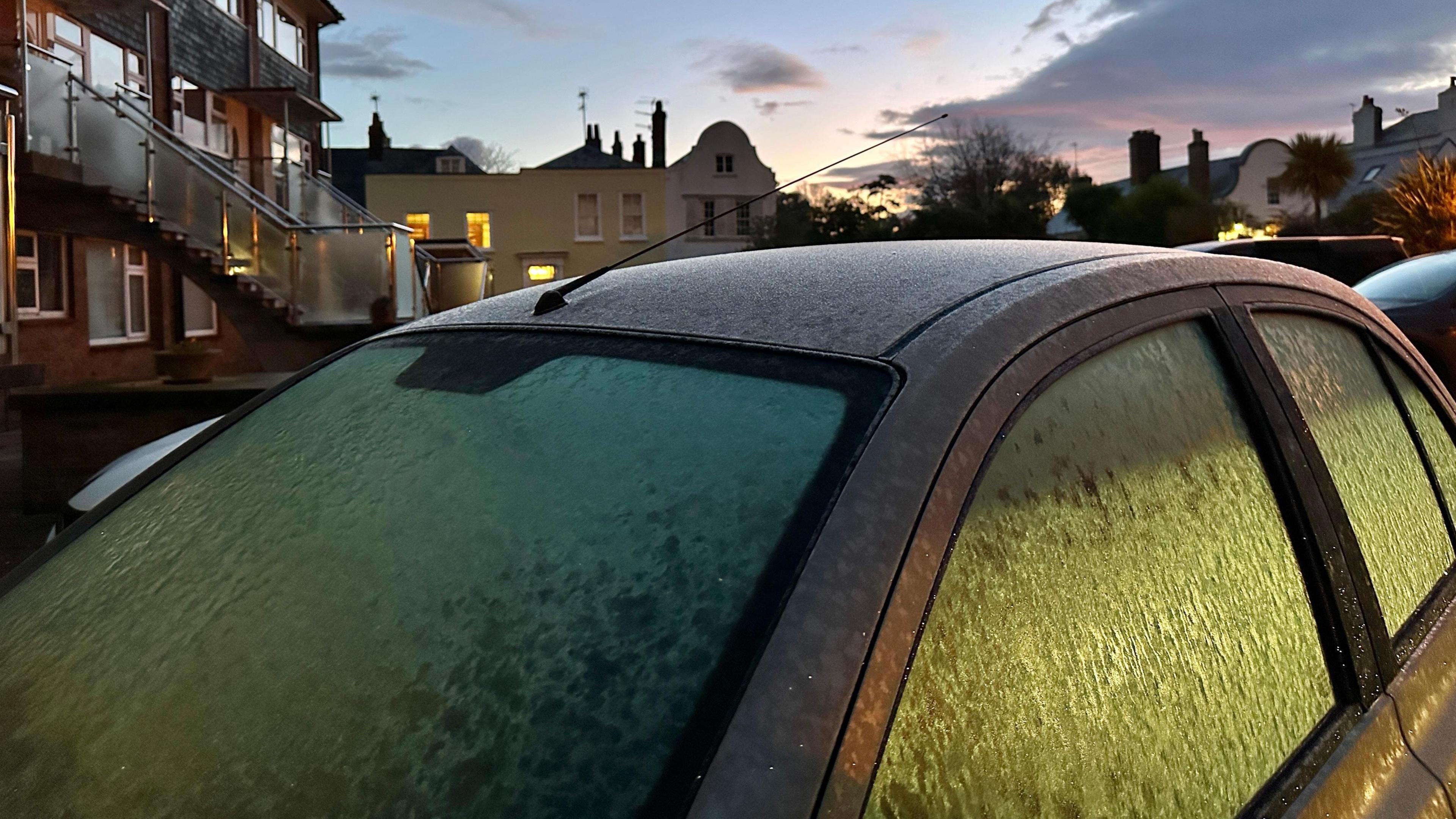 A car is seen with its windows covered in ice, houses can be seen in the background 