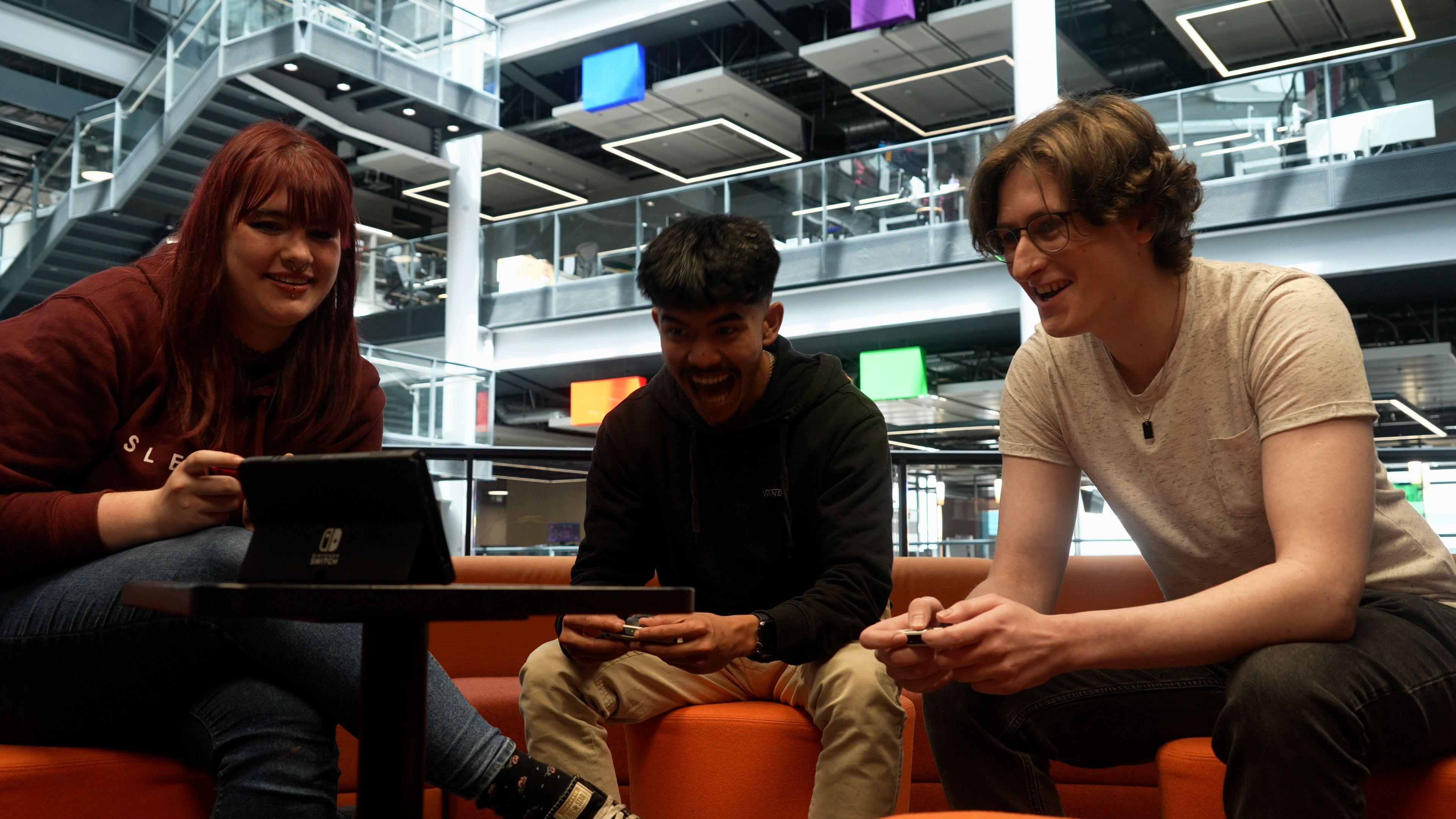 Kady, Dirk and Nicholas sit on orange sofas playing a multiplayer game together on Nintendo Switch. They're laughing and smiling during a tense race.