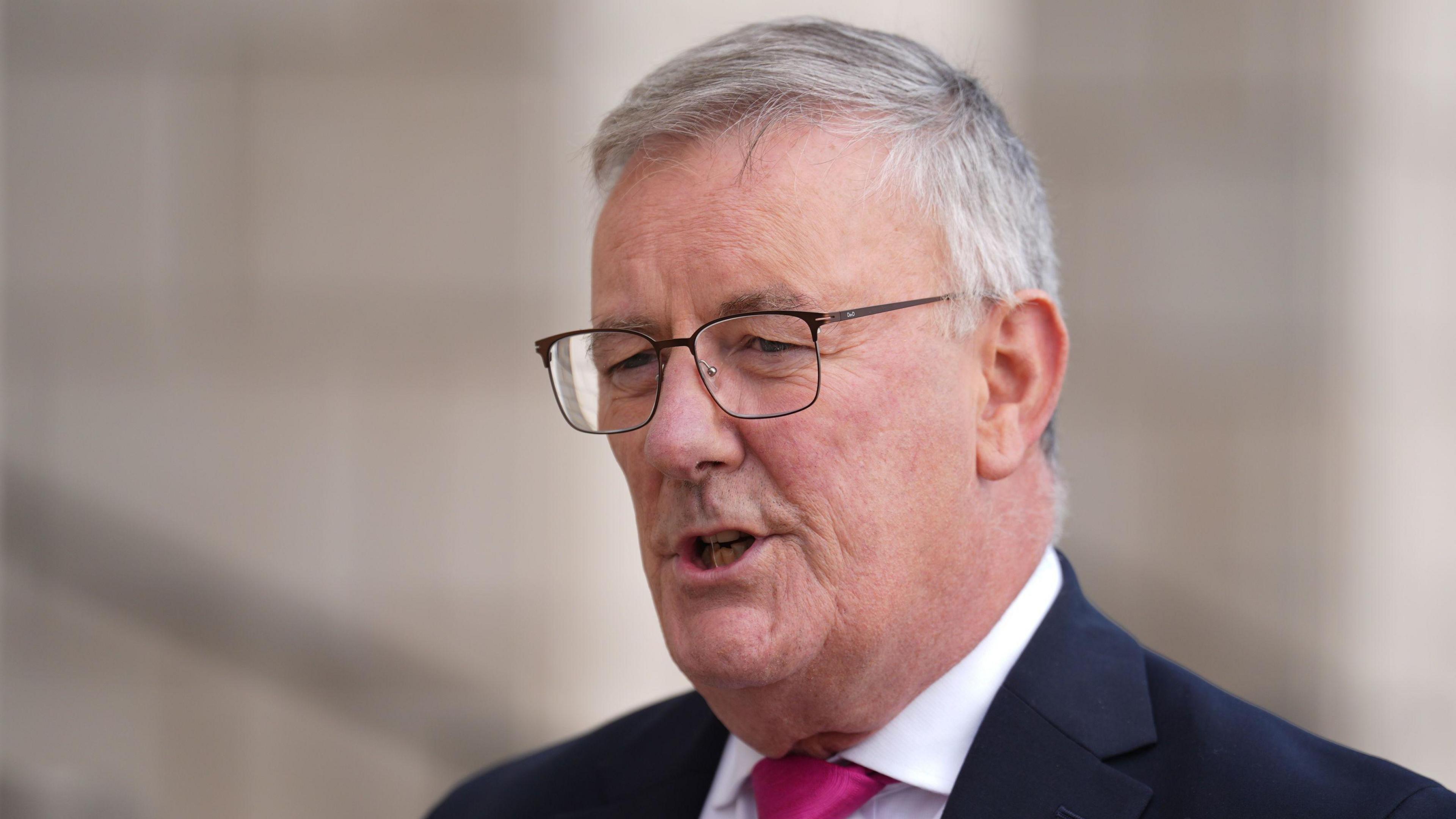 Mike Nesbitt has short grey hair and metal square glasses. He is speaking. He is wearing a white shirt, pink tie and a black blazer.