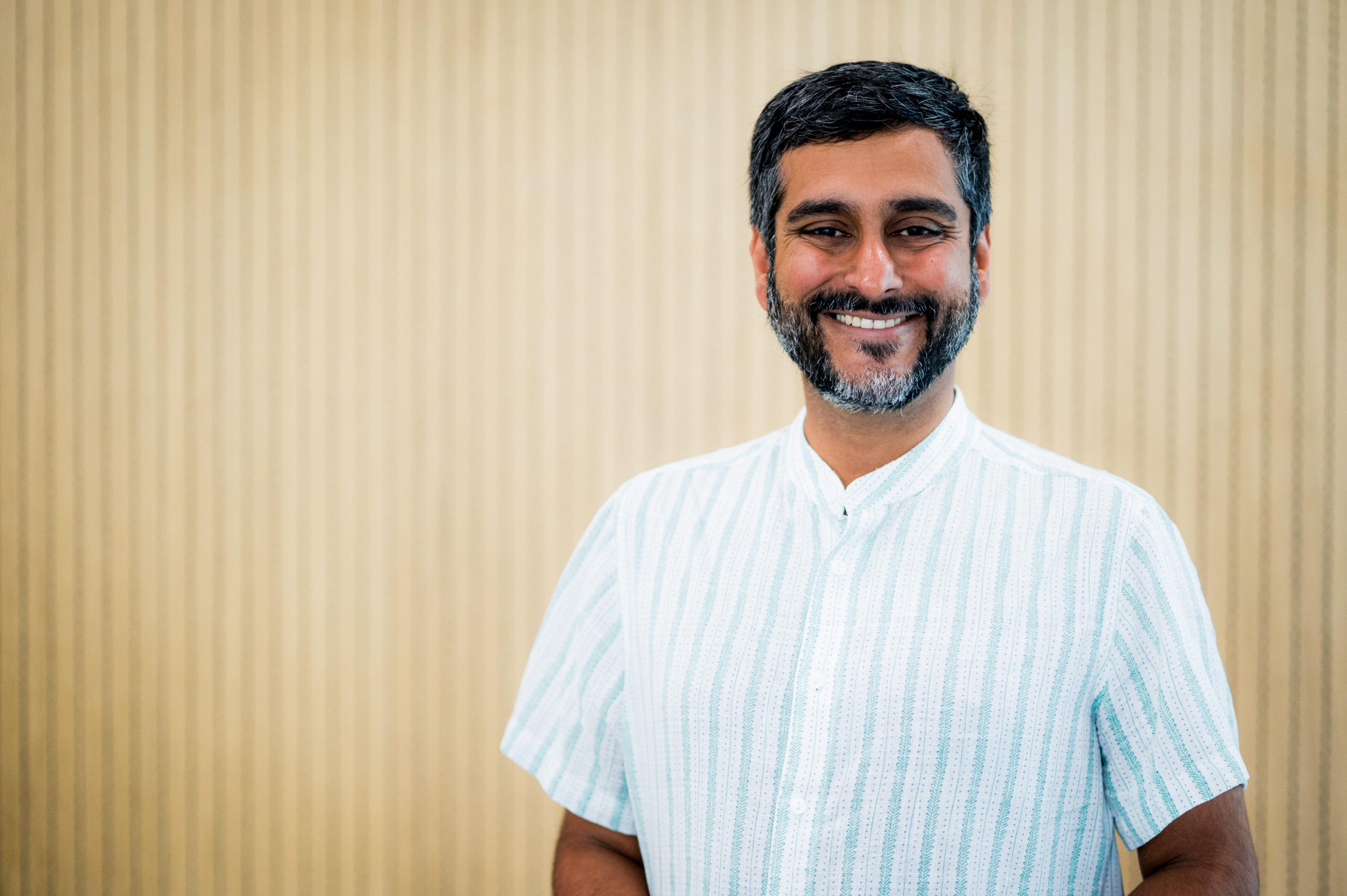 Dr Vivek Nityananda is standing and smiling at the camera. He has short black hair and a black/ grey beard.