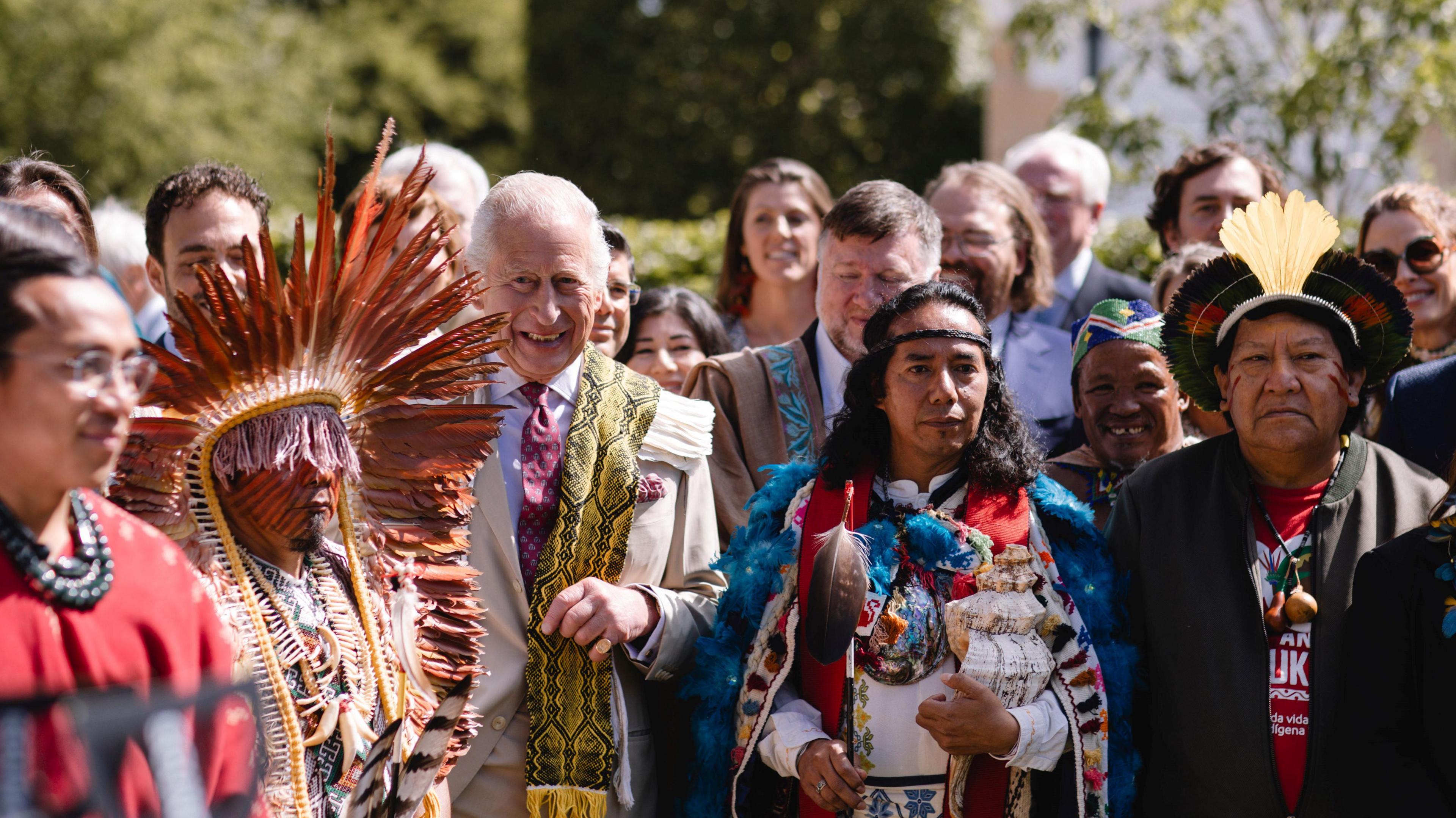 King Charles and delegates at the Harmony Summit in Highgrove. He is draped in a yellow and black patterned scarf and standing between several men in traditional dress including one in a feathered headdress