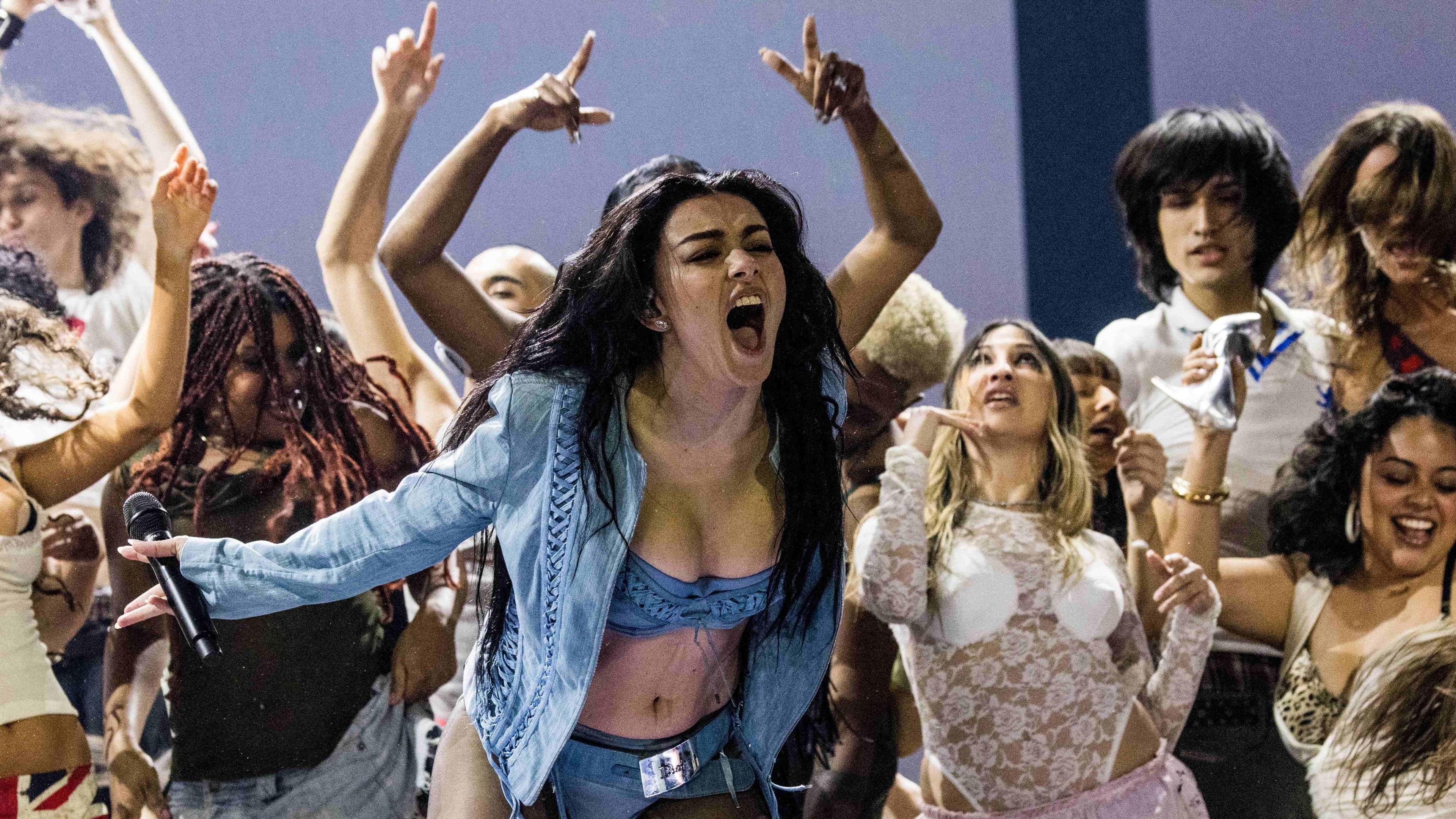 Charli XCX performing on stage at the Grammy's. The singer wears a denim look made up of a bra top and pants under a long-sleeved matching jacket. She sings into the crowd, her long dark hair worn loose. She's surrounded by dancers. 