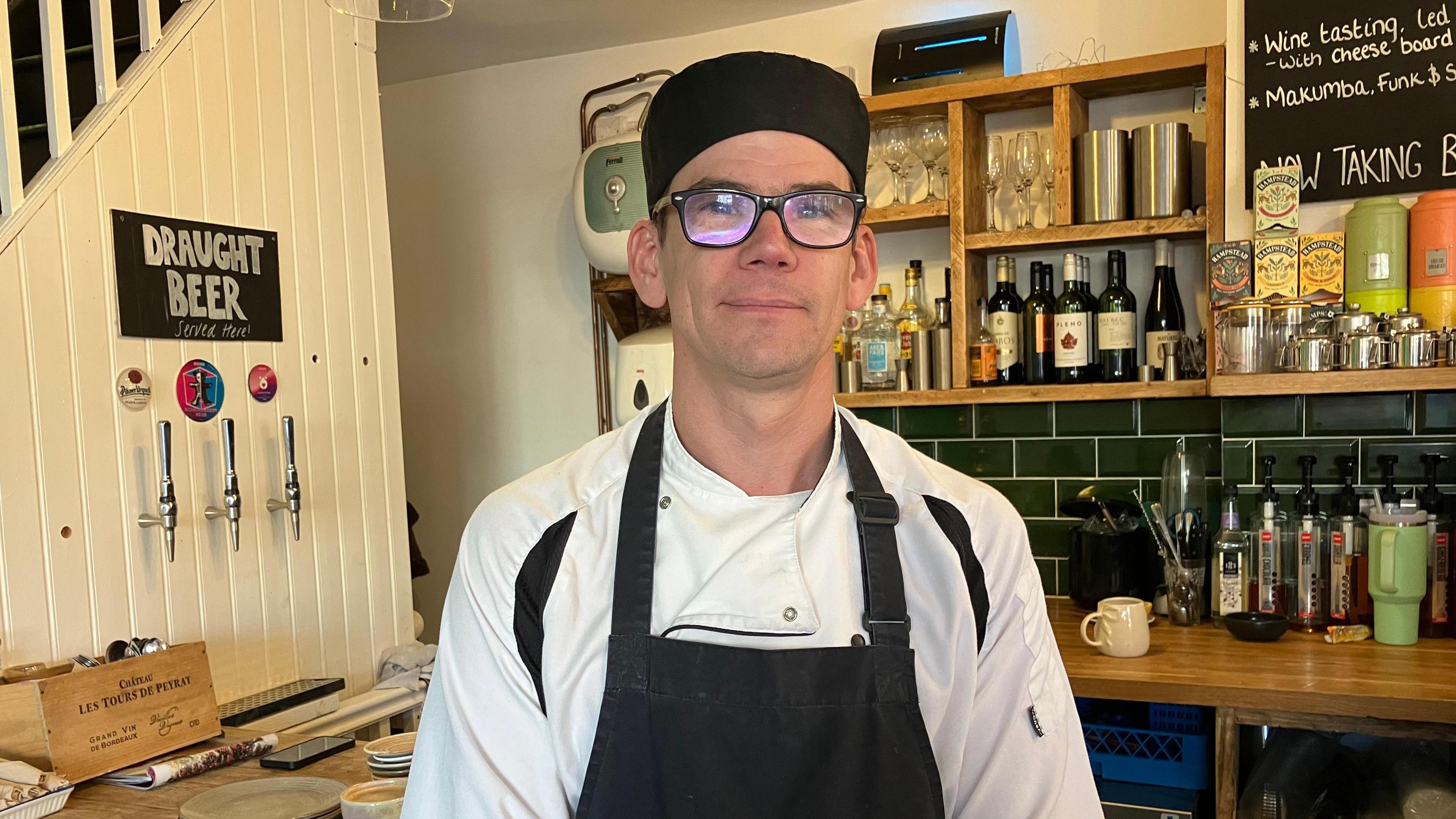 Jolyon Iles standing behind the counter of The Beech Tree wearing a chef's hat, apron and chef white tunic and is looking into the camera. Bottles of wine, spirits and syrups are on counters and shelves behind him. Beer pumps can be seen on wall panels.