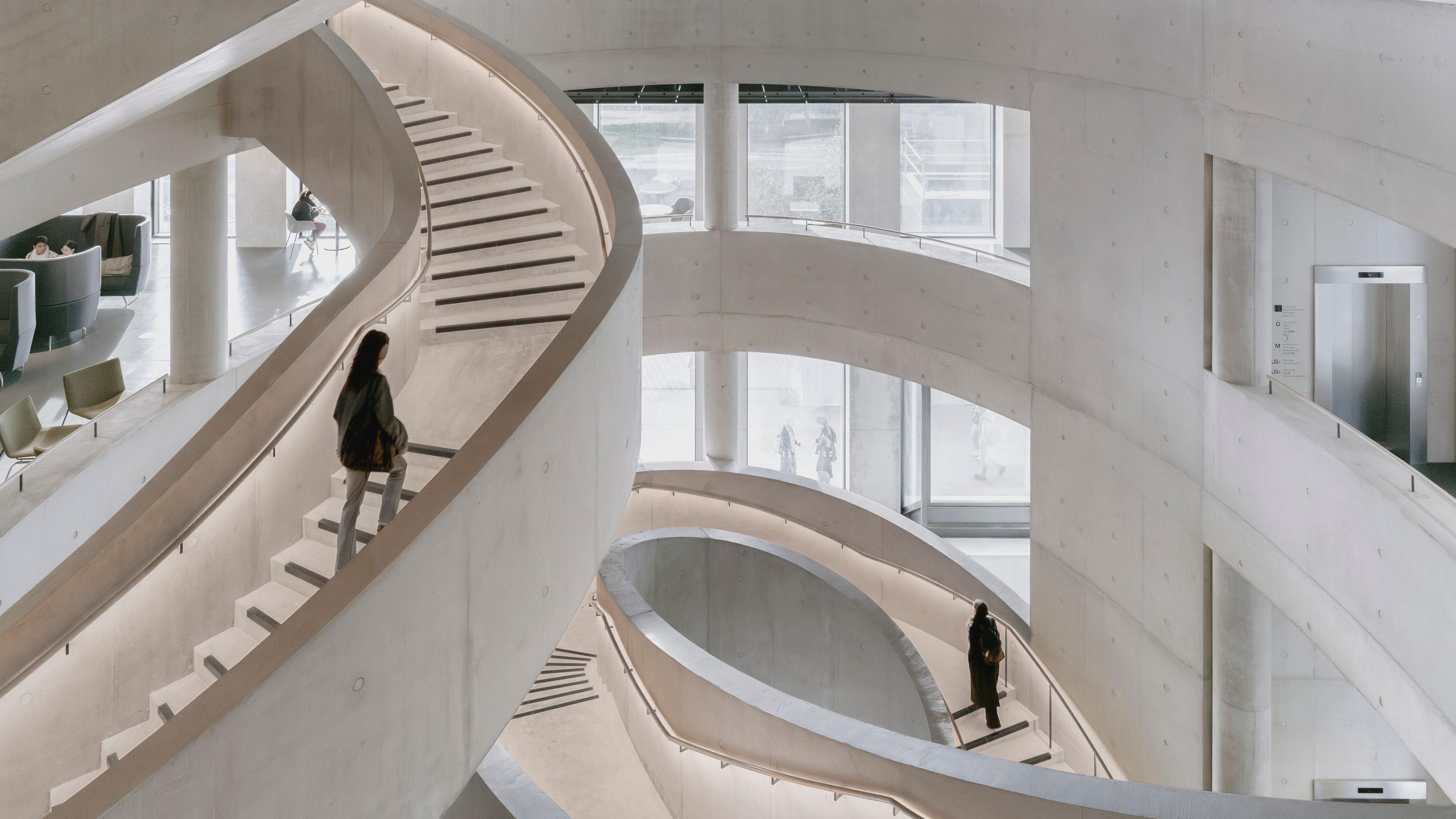 Winding, white concrete staircase. One person is walking up one and another is in the distance walking up another staircase. The building's interior also has a concrete-look and is white. 