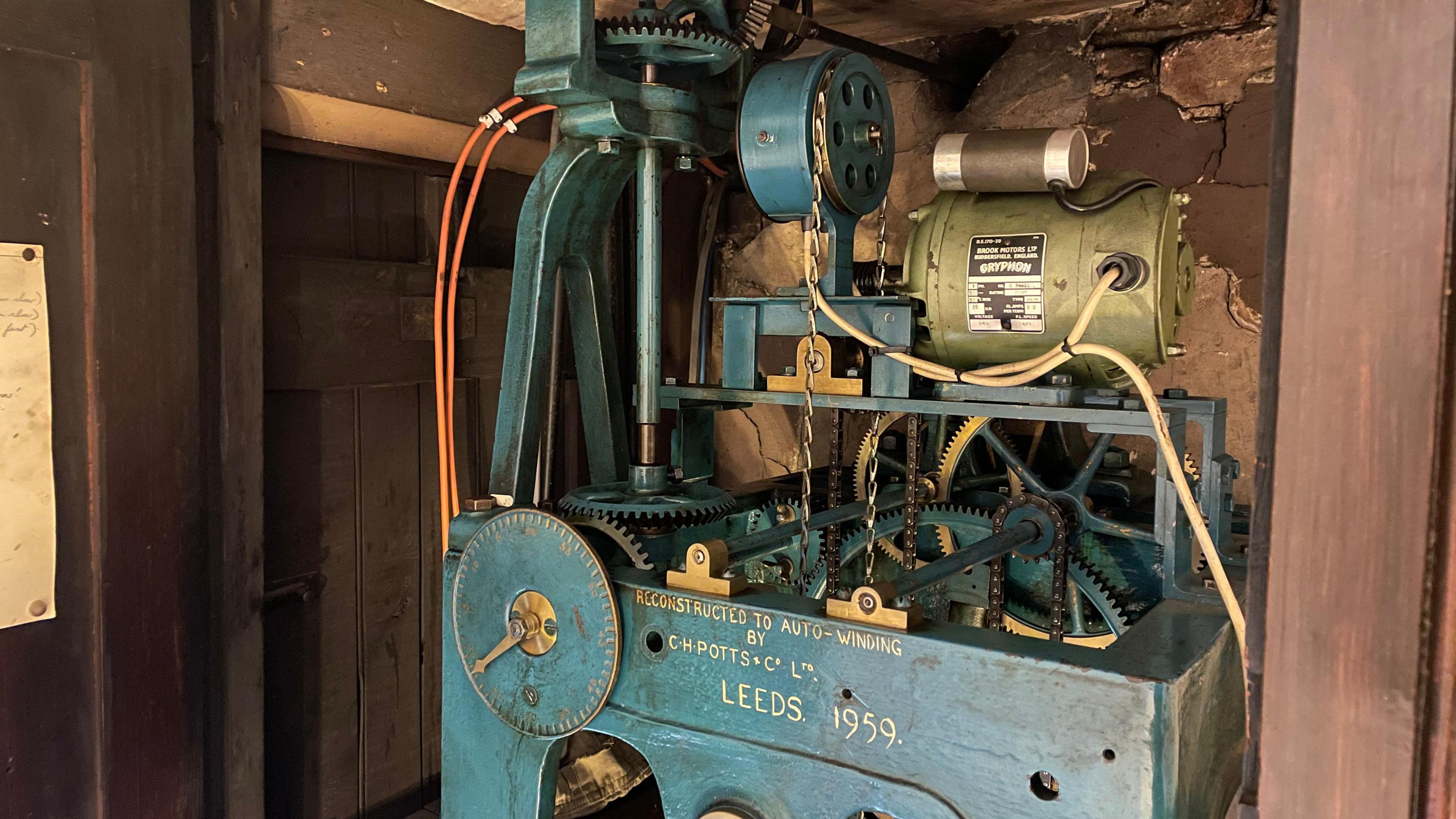 A blue painted clock mechanism. There are chains and pulleys and a green coloured motor with cables coming from it.