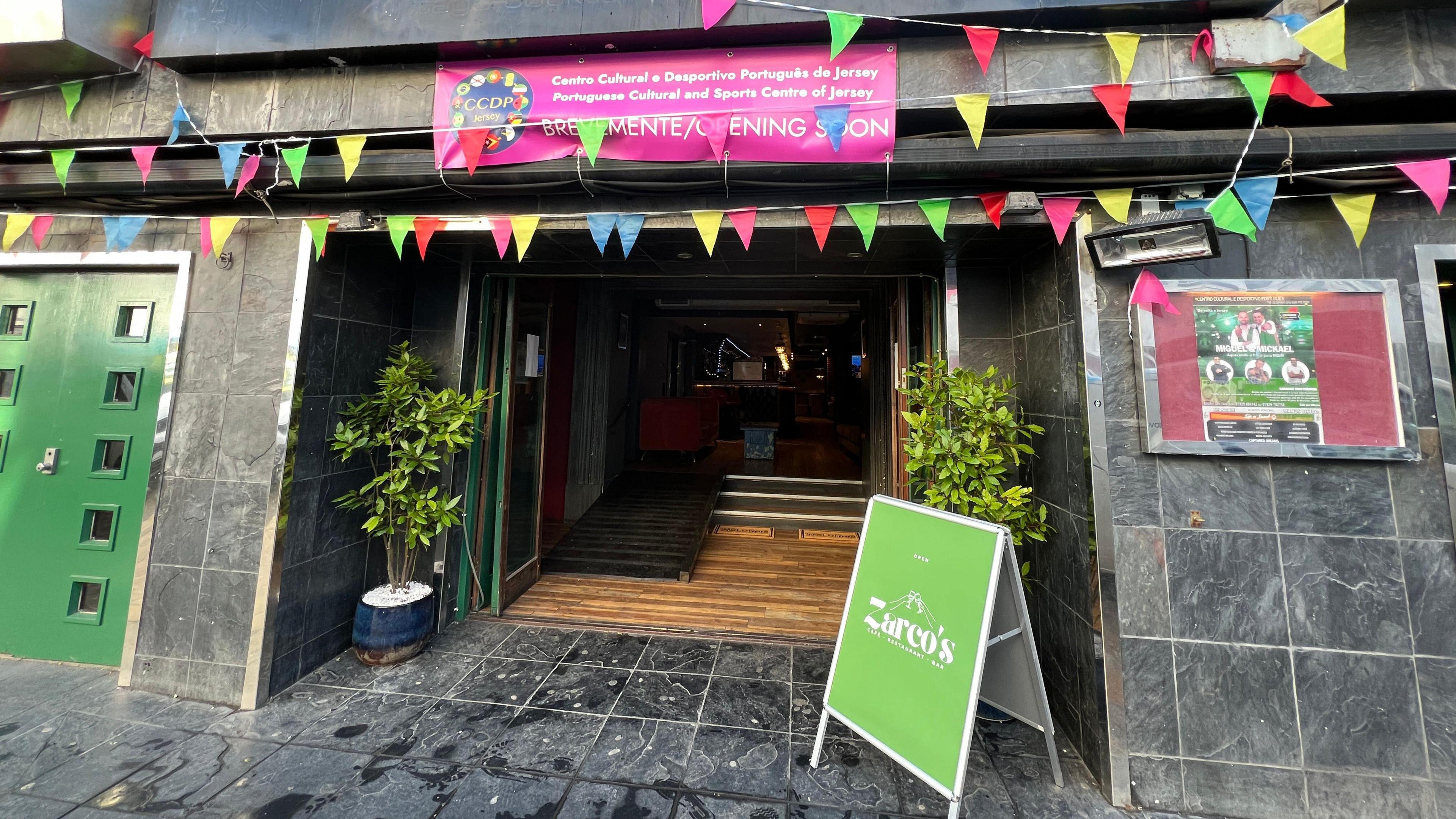 Colourful bunting flags hang over an open doorway which has steps and a mobility ramp going into the cultural hun. There's a green billboard sign outside with two plants either side of the door. There's a pink banner up over the doorway which has the CCDP logo on it.