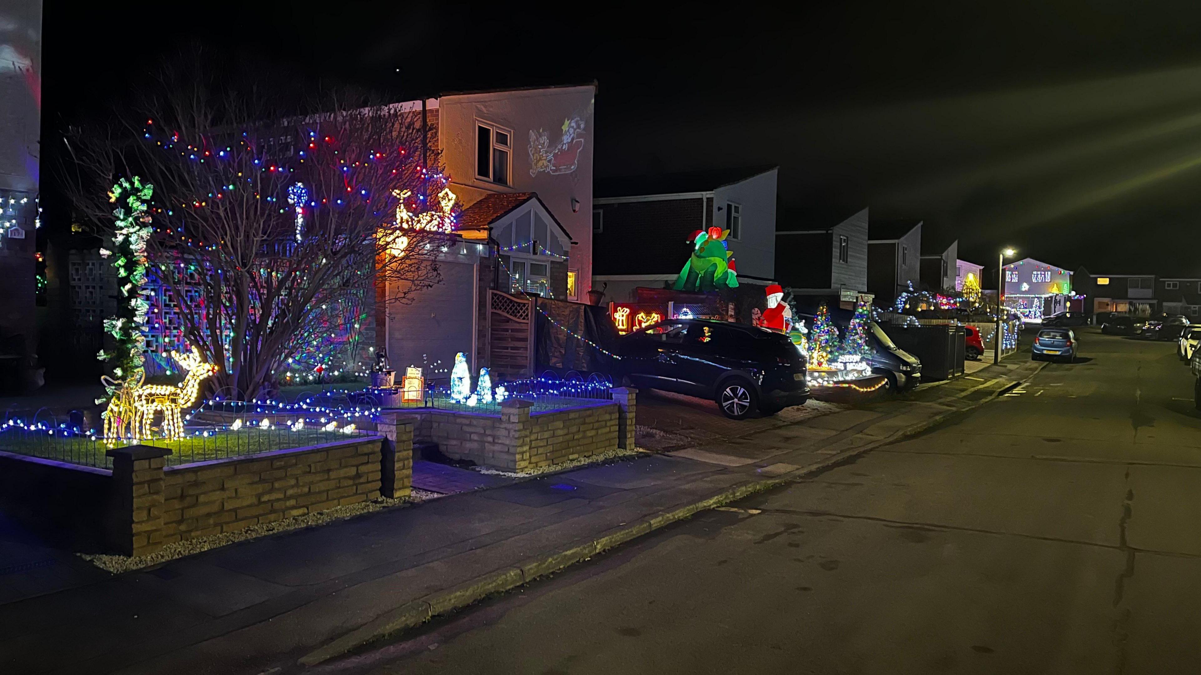 A line of houses along the street are all individually lit up with lights, reindeer figurines, Christmas trees and other festive characters. The pavements are empty, but the houses are glowing.