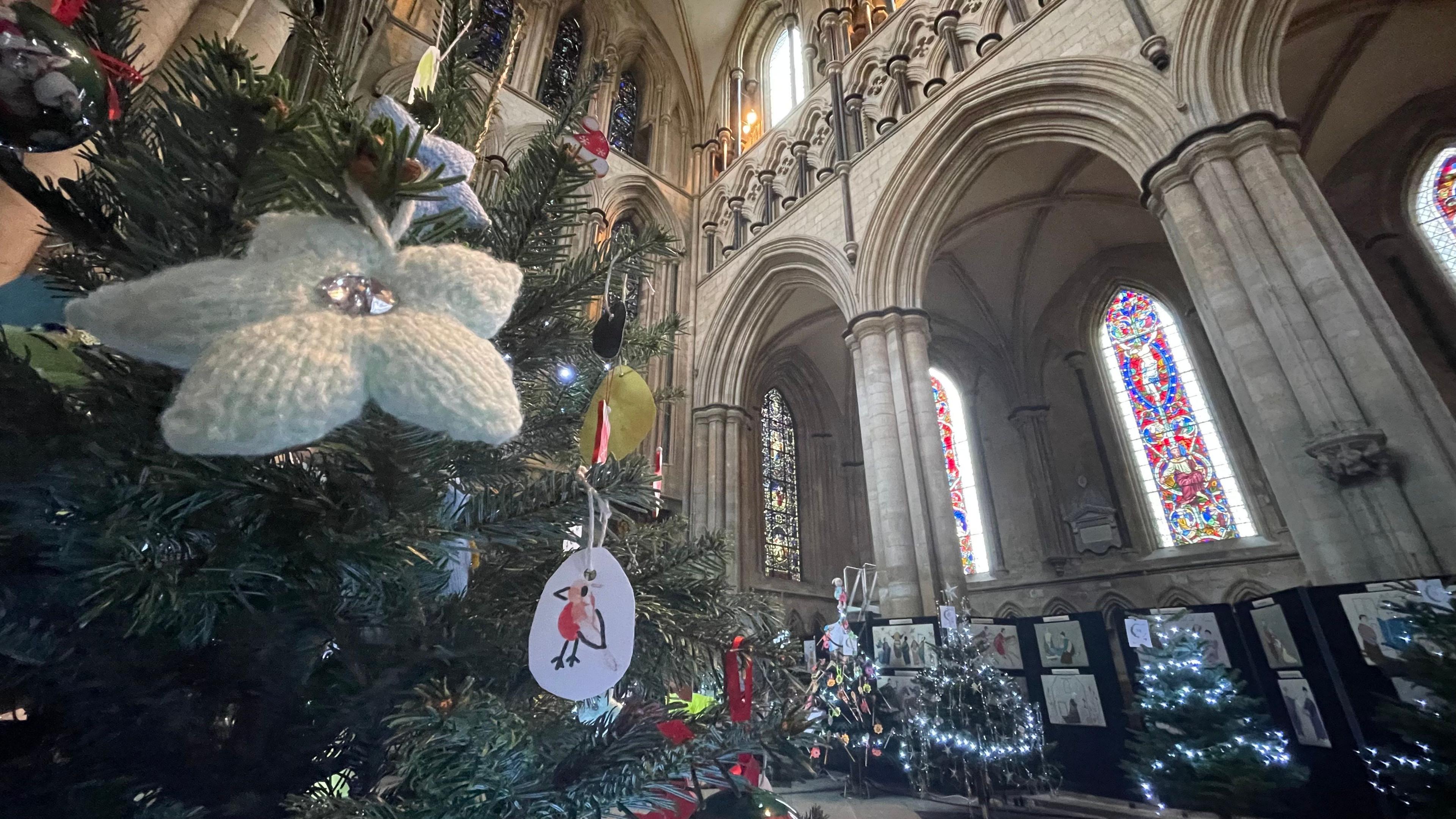 In the foreground, a white knitted Christmas tree decoration with a bead at its centre, hangs on a fir tree next to a decoration with a drawing of a robin. In the background are four other decorated Christmas trees. The came is angled upwards to show the archways and stained glass windows of beverley Minster