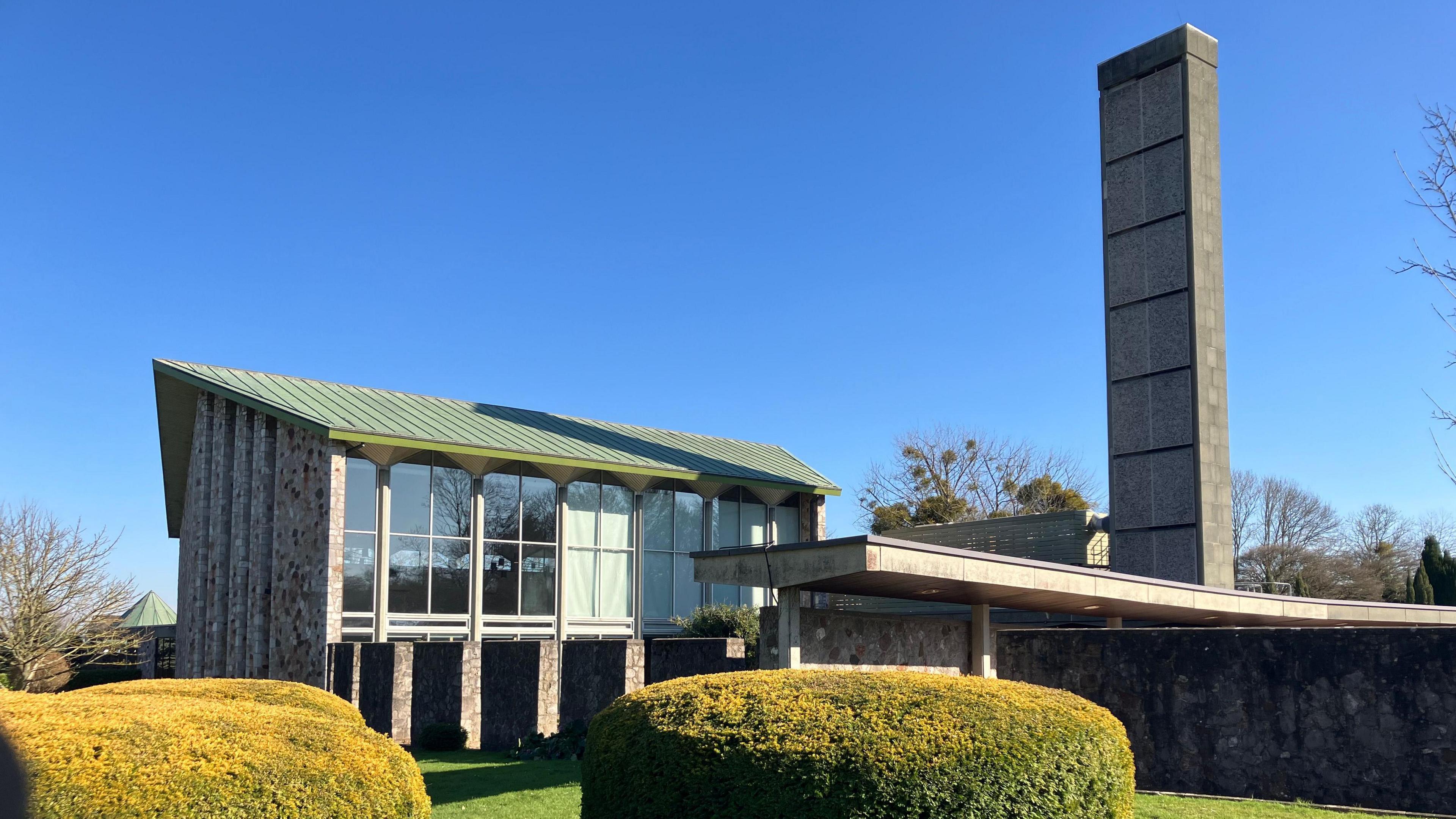 The picture shows Taunton crematorium on a sunny day.  The image shows the main building, walkway and two bushes in the foreground.