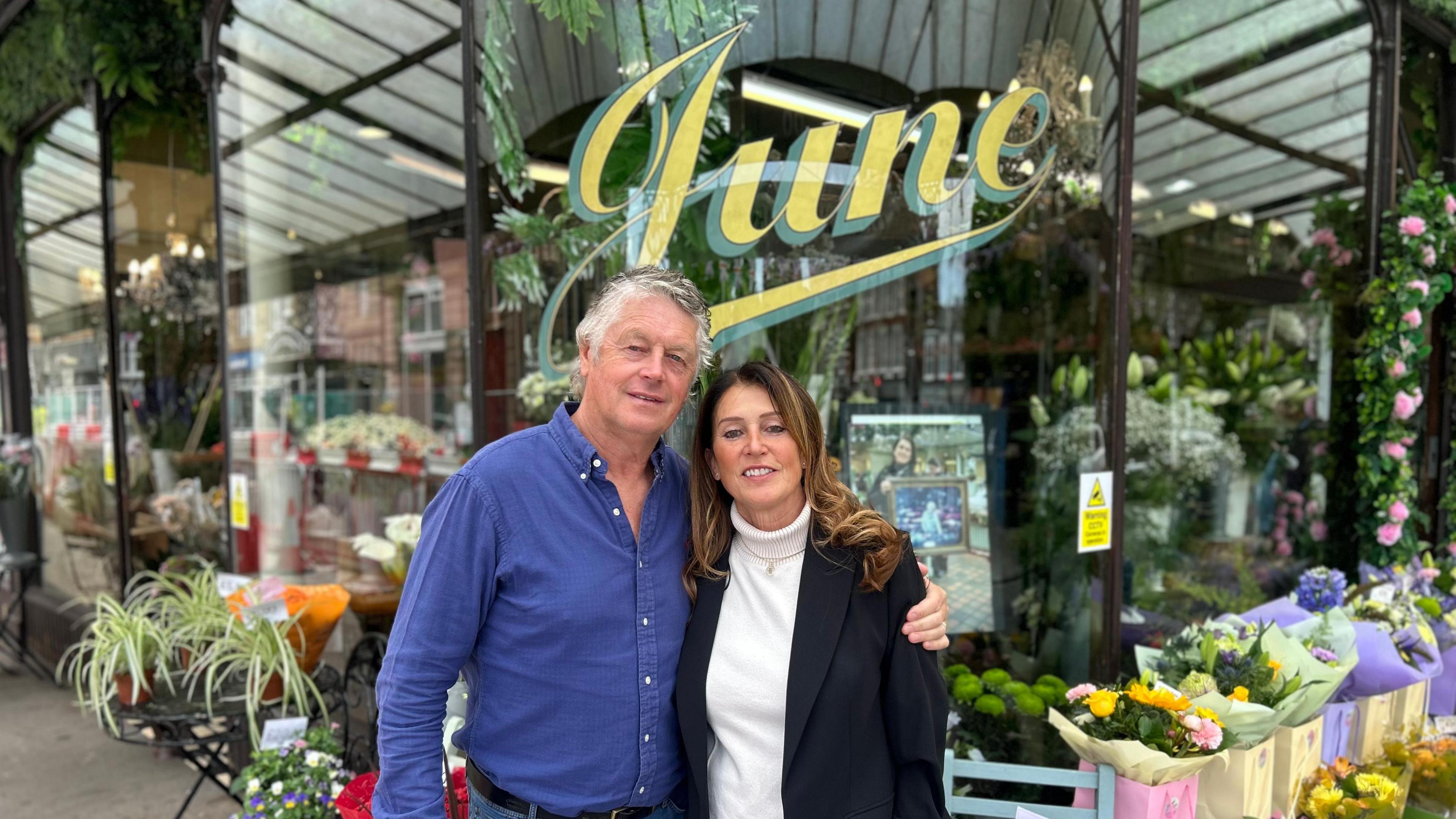Peter and Liz outside the florist - which has June in italic lettering on glass windows - they are surrounded by flowers and plants. Peter has a blue shirt and Liz is wearing a white polo neck and black jacket.