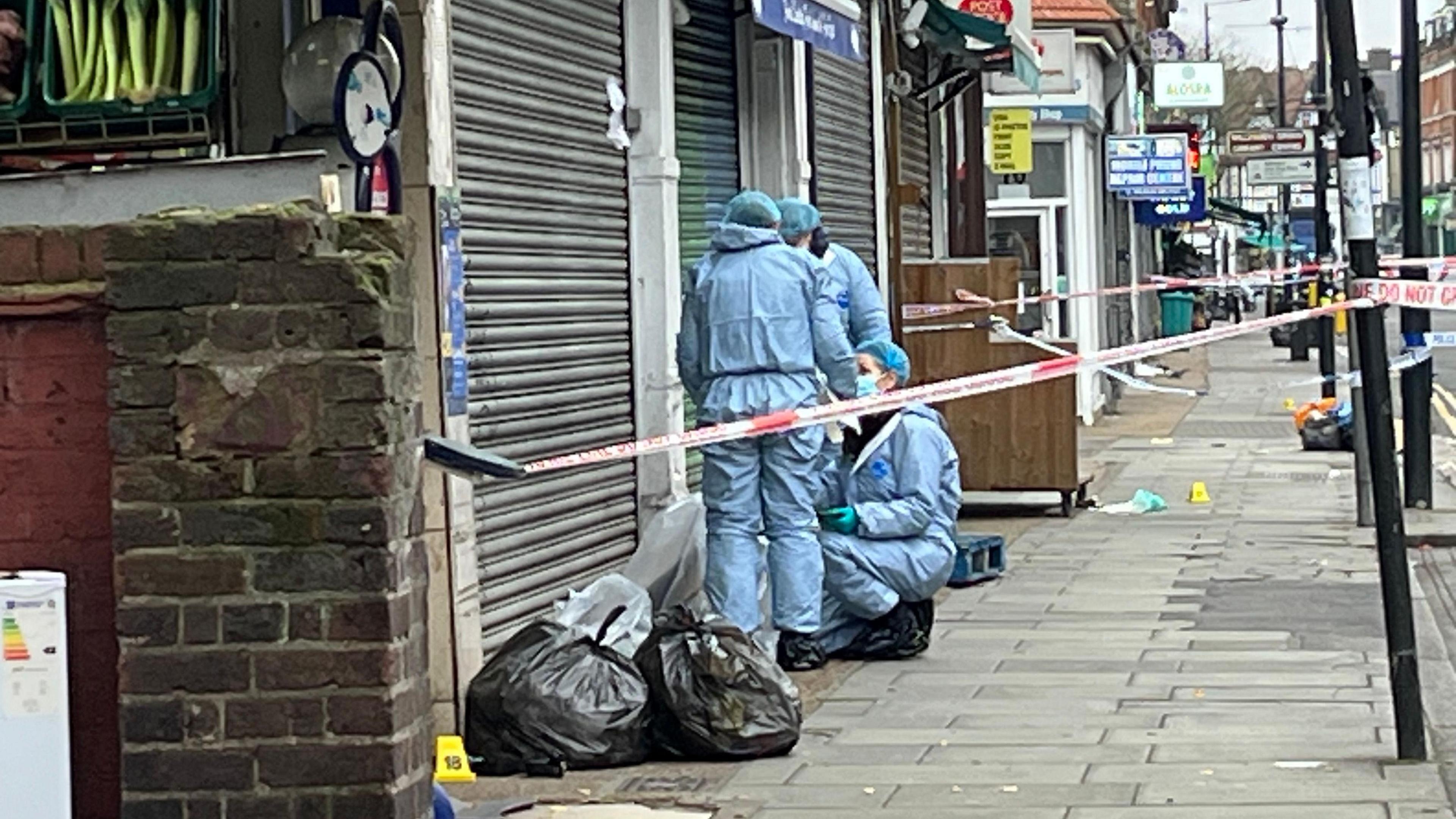 Three people in blue all-in-one suits, gloves, hair covers and masks work within a police cordon outside a shop with shutters pulled down and next to rubbish bags on a high street.