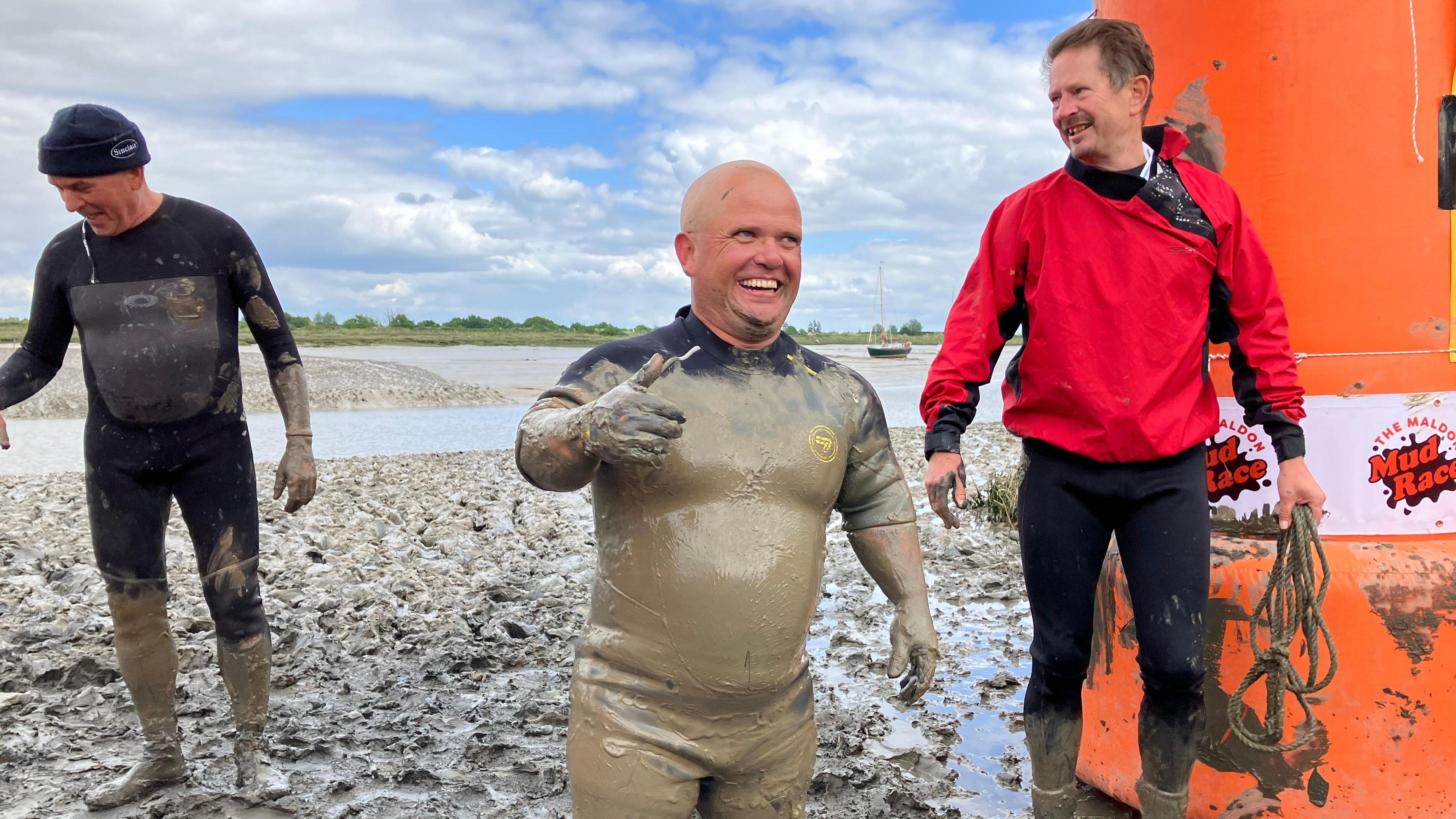 A man in a wetsuit coated head to toe in mud gives a thumbs up at the finish line
