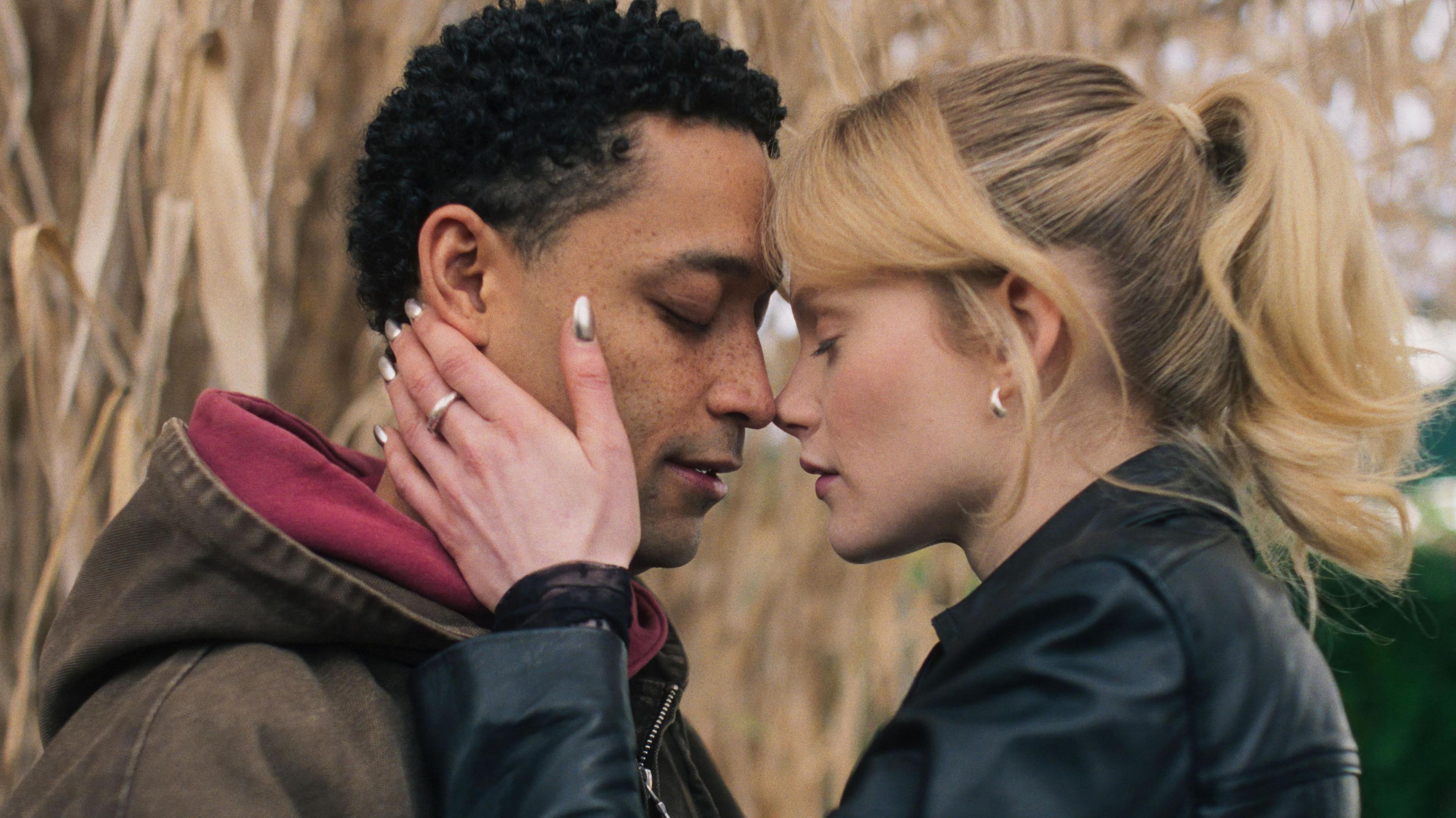 A woman wearing a black leather jacket puts her hands on the face of a man wearing a grey jacket and red hoodie, as if she's about to kiss him
