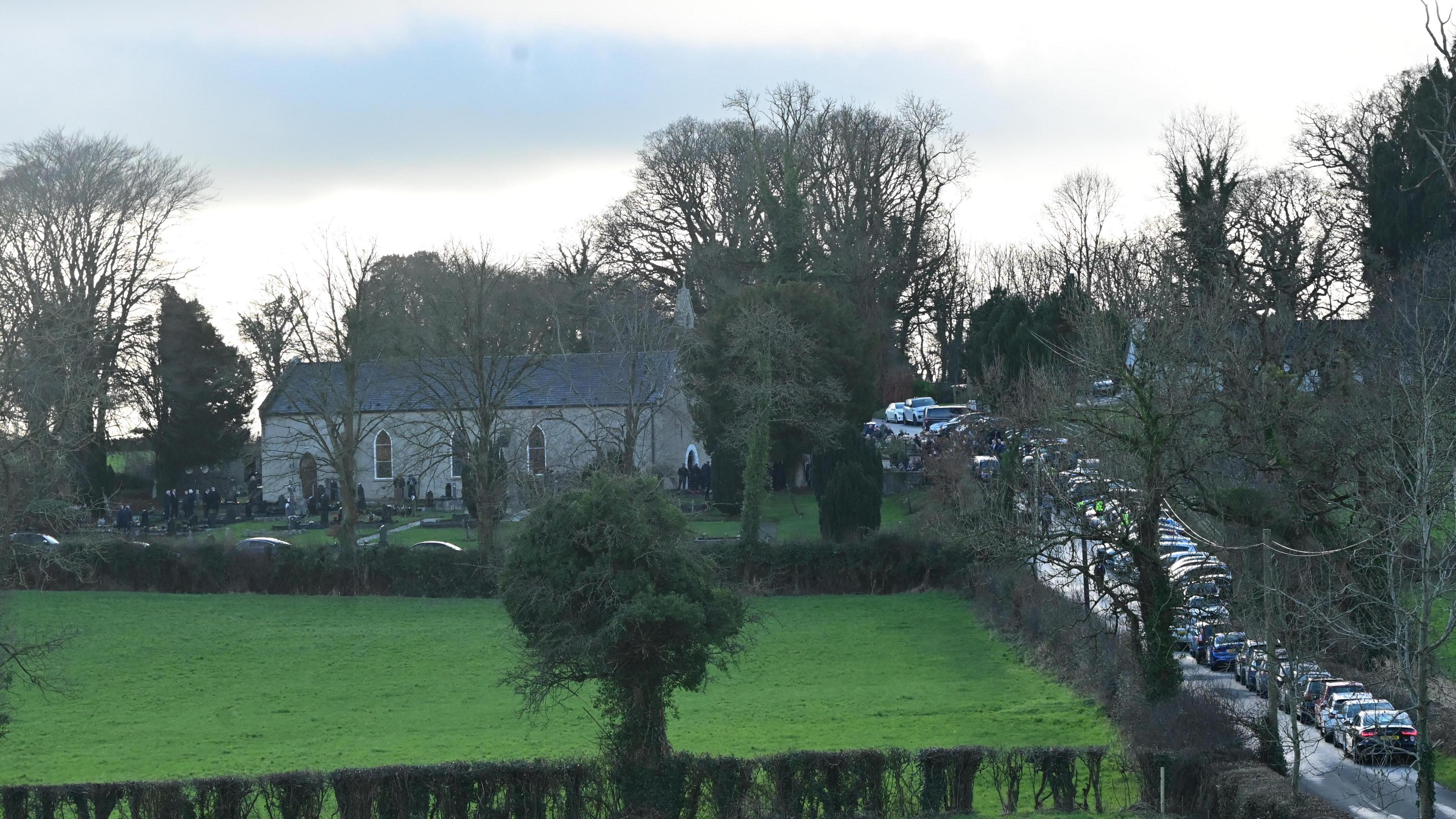 A far away shot of the church where the funeral took place. Fields roll from a stone church. Cars are parked on the country roads. 