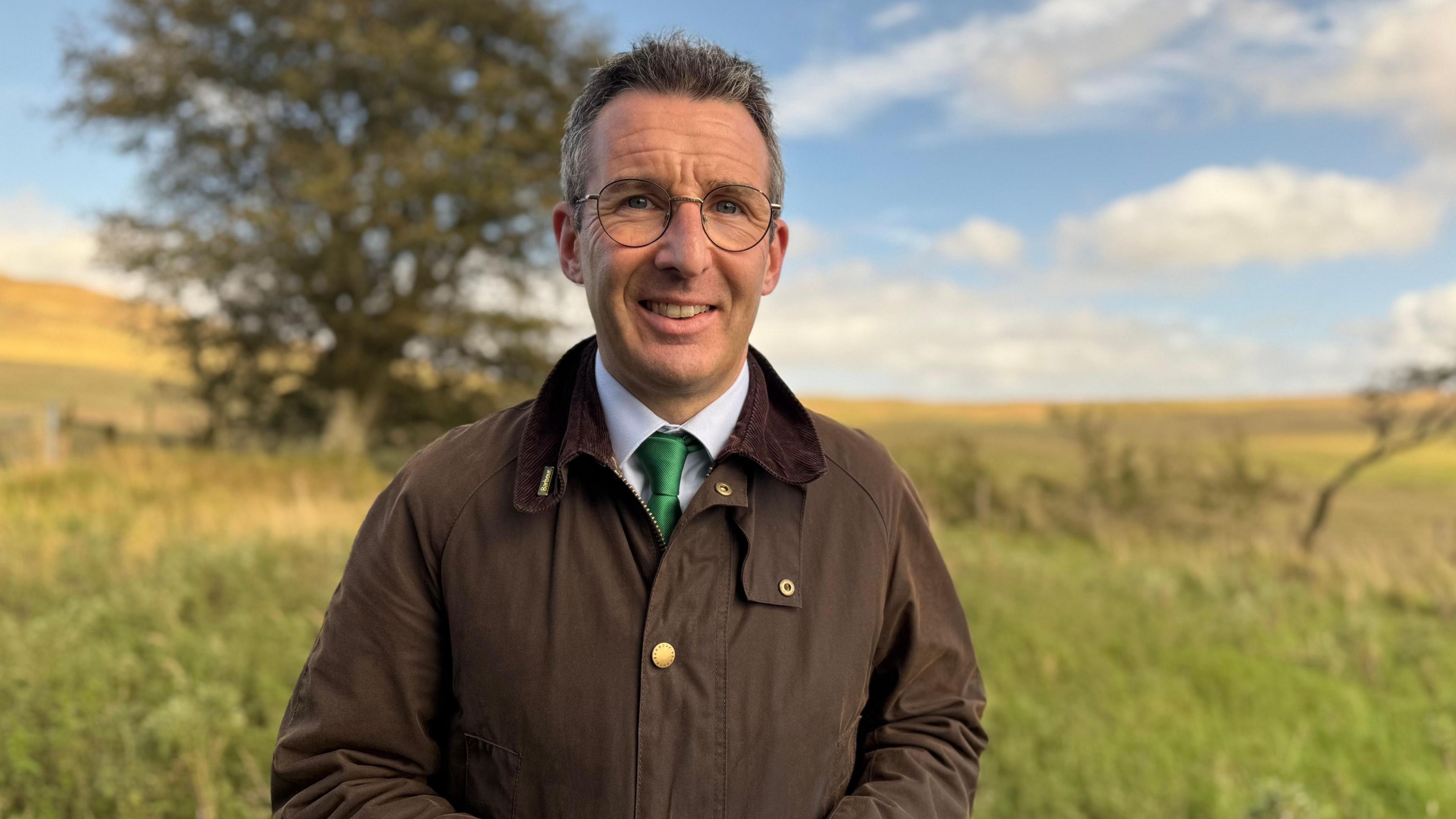 Andrew Muir standing in a field on Divis Mountain on a sunny day. He has short, dark, greying hair and glasses. He is wearing a brown wax jacket, a blue shirt and a green tie. There is grassland and a large tree behind him.