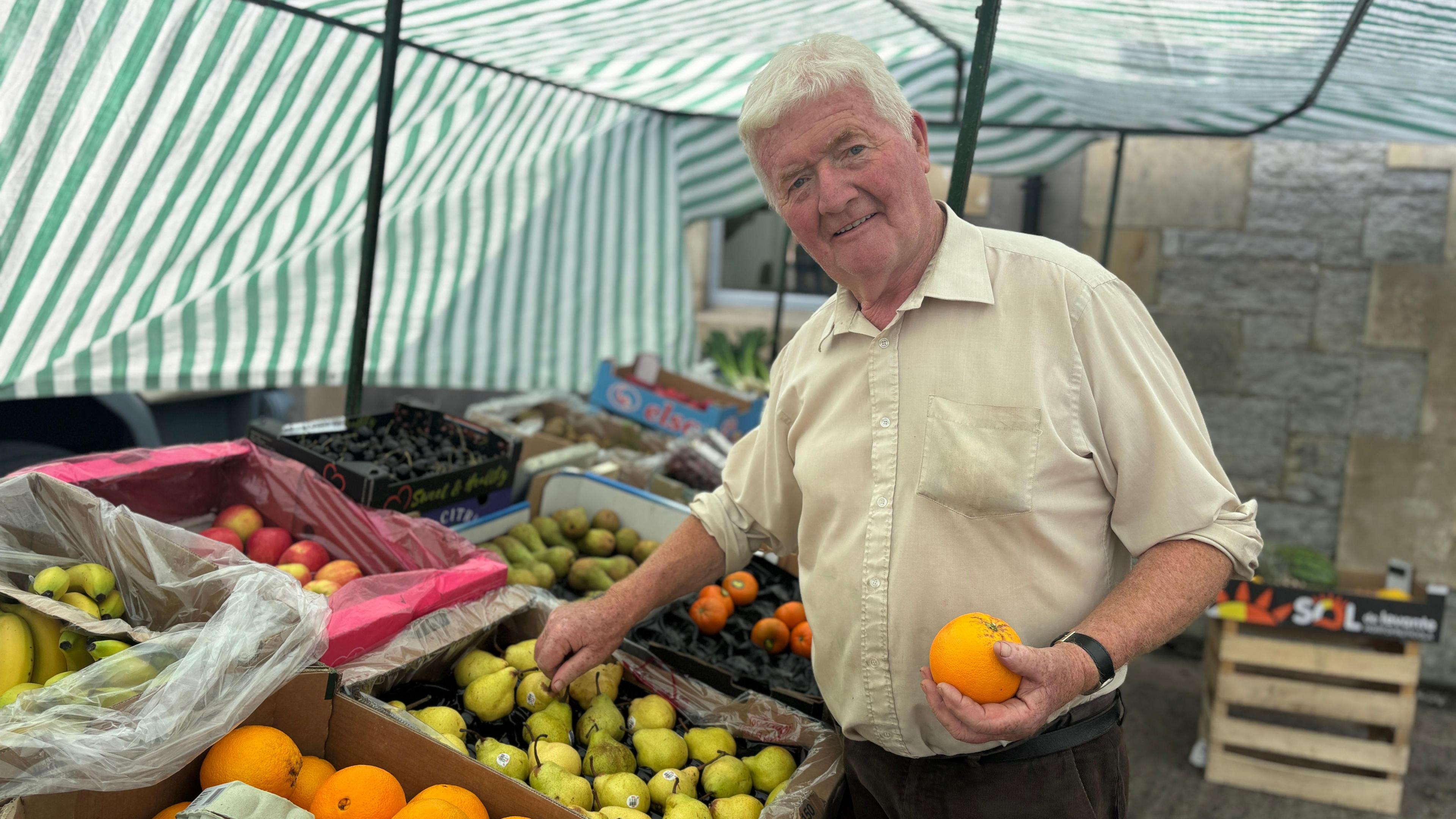 A man stands at an outdoor market stall. There are boxes of fruit and veg to his left. He is holding a large orange in one hand. He has grey hair, a smile and is wearing a light yellow shirt. Green and white striped awning covers the area where he is standing.