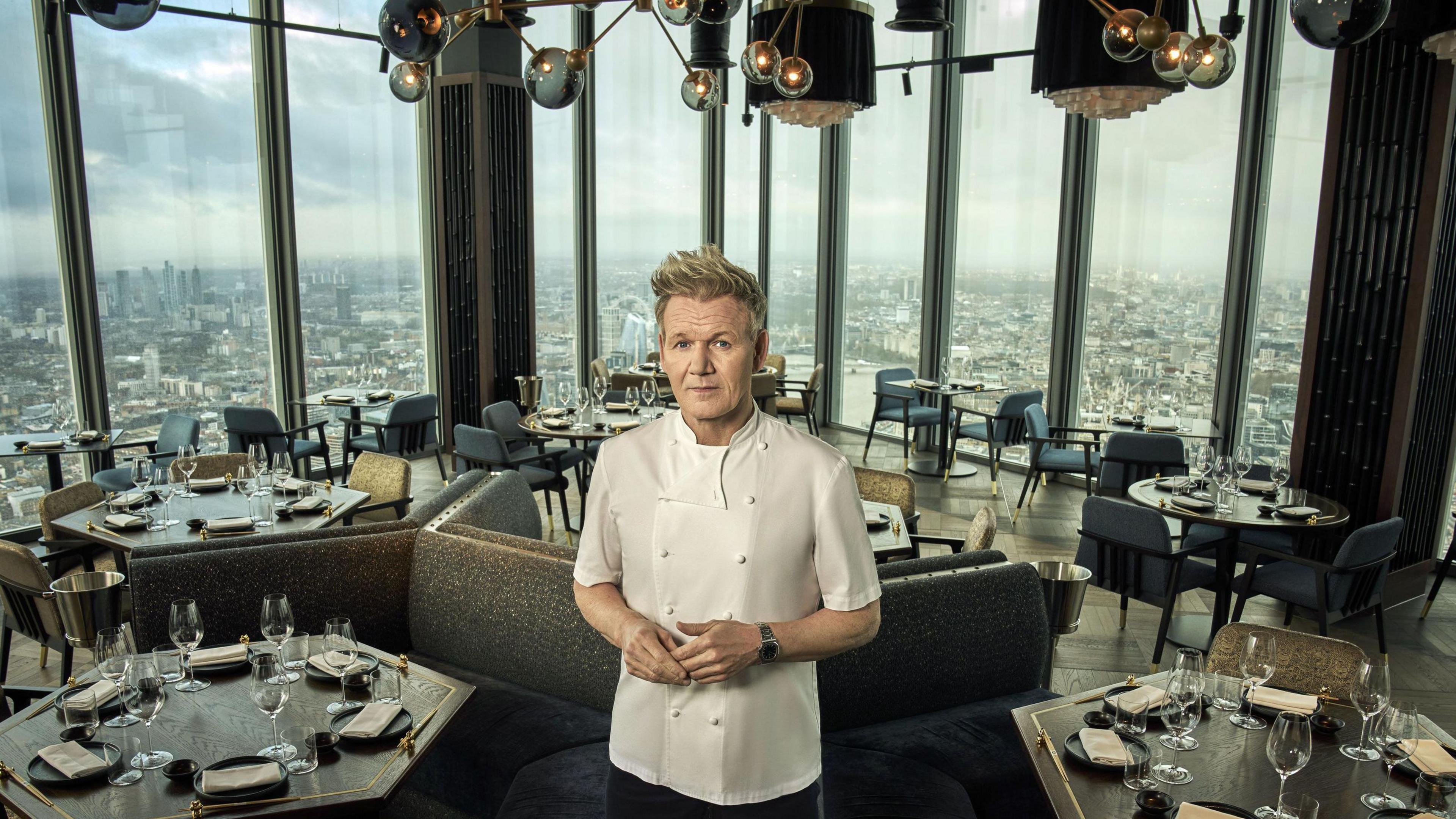 Gordon Ramsay stands in the middle of a high-rise restaurant with large windows showing a panoramic view of London. The restaurant has dark wooden tables set with wine glasses, plates, and napkins, surrounded by plush chairs and booths. The lighting fixtures above have a modern, gold-toned design.