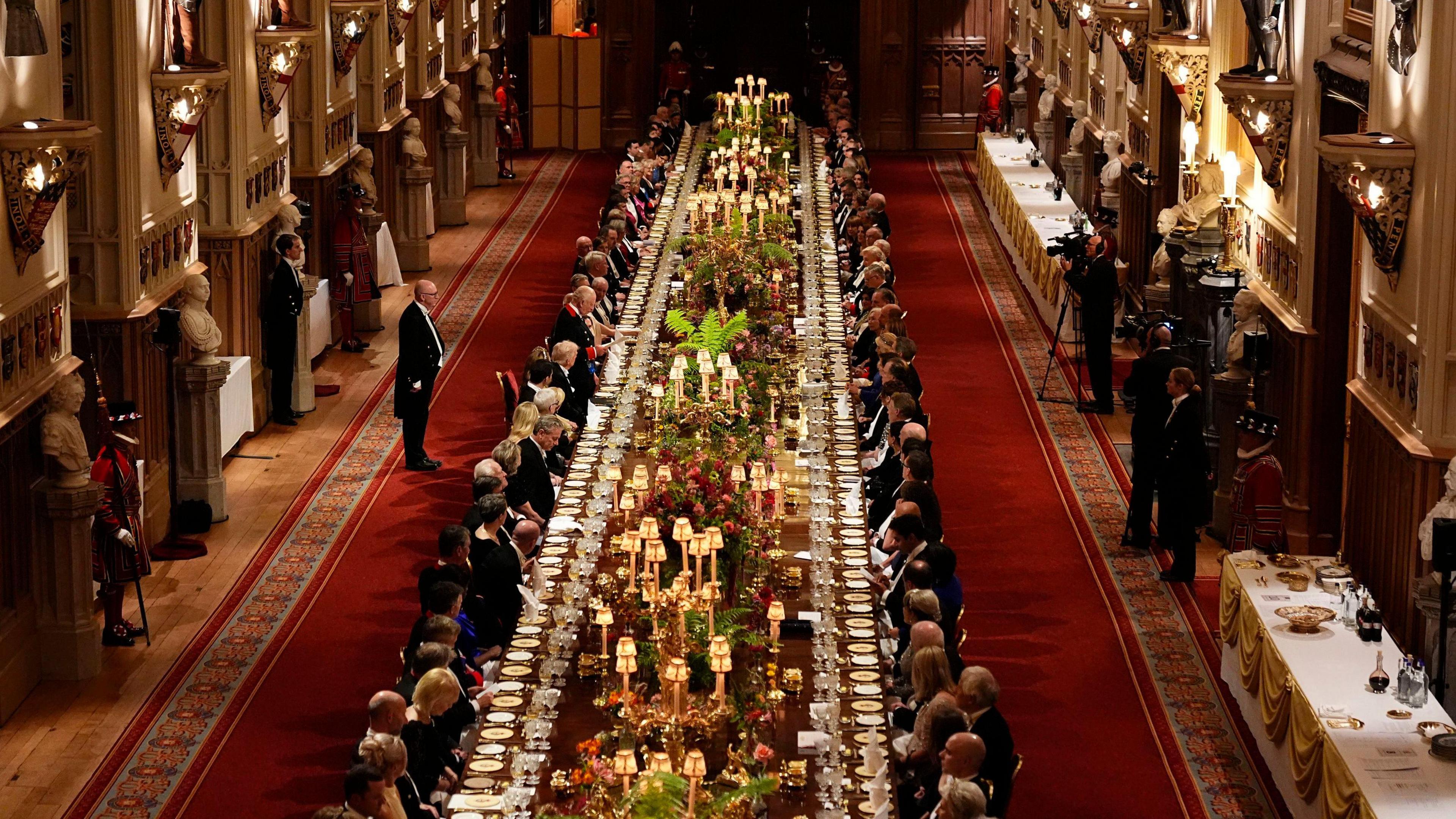 A long dining table with dignitaries seated down either side is seen in a banquet hall, with staff and press against the walls.